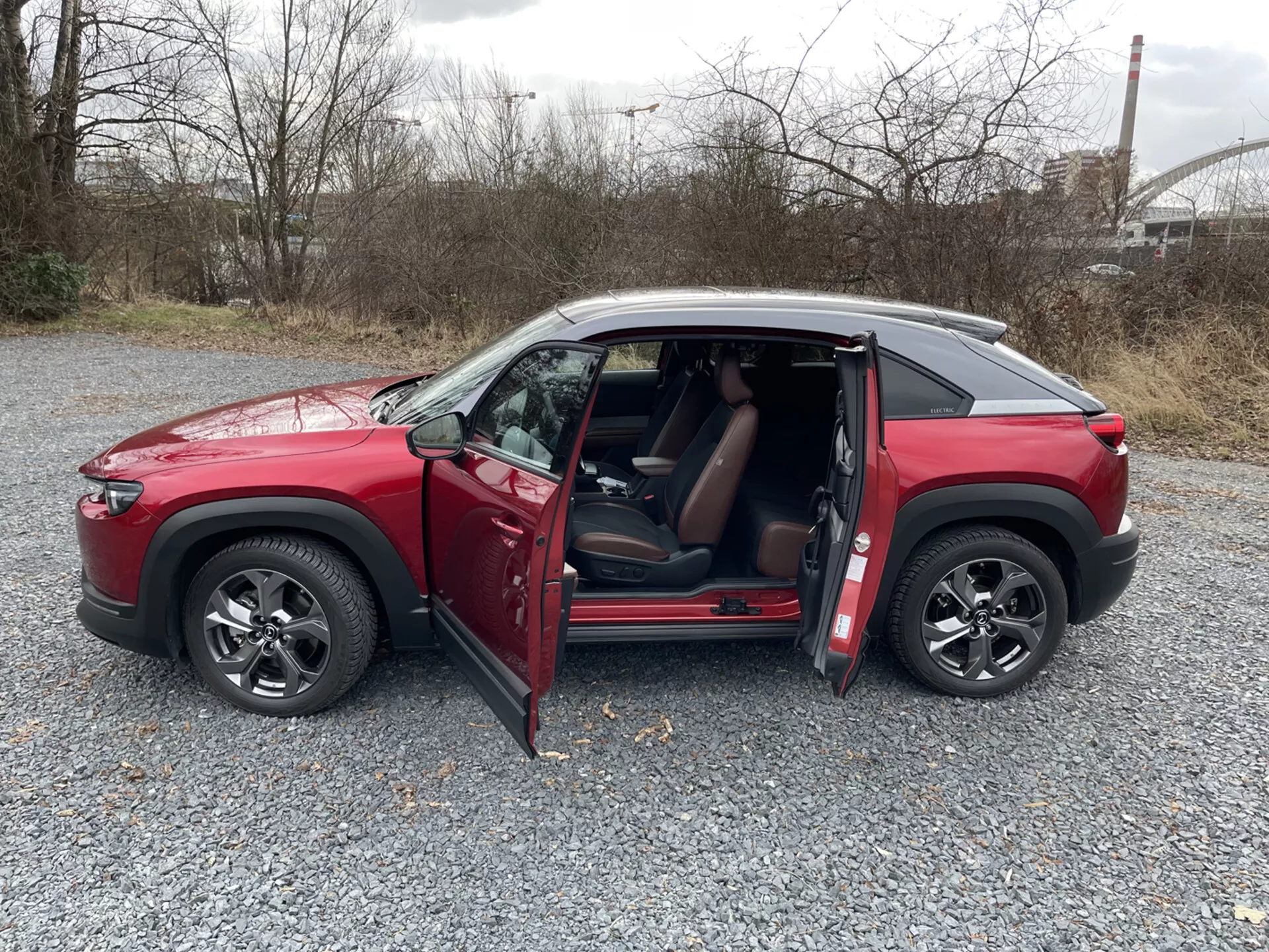 Profilo laterale SUV rosso Mazda MX-30 2020 con quattro porte e porte lato conducente anteriore e passeggero posteriore aperte, parcheggiato su ghiaia sotto illuminazione nuvolosa, con ruote in lega e uno sguardo al suo interno, presumibilmente rivestito in pelle, con tecnologia visibile sul cruscotto.