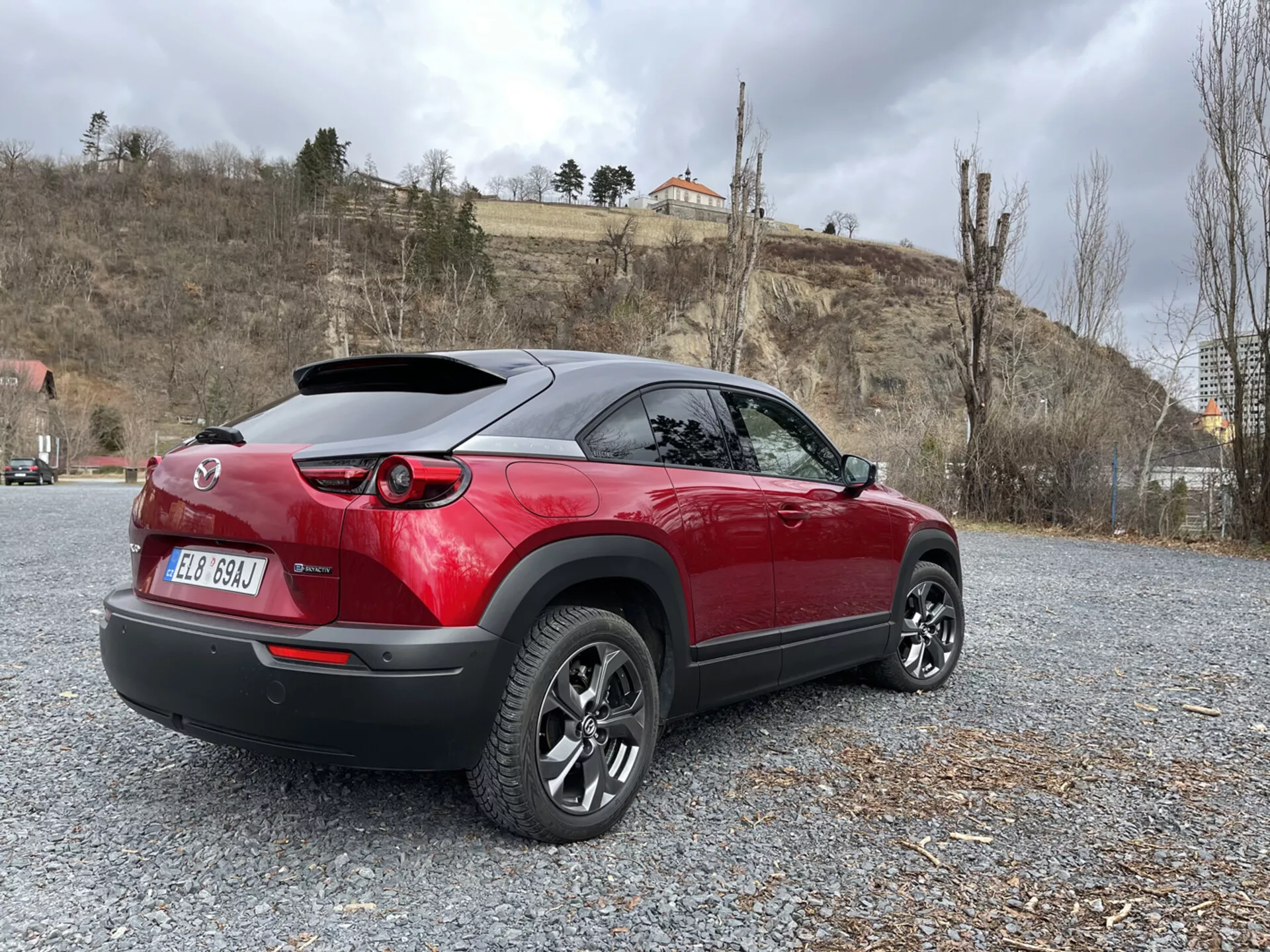 Crossover moderno Mazda MX-30 2020 parcheggiato su ghiaia, che mostra il suo design sportivo bicolore rosso e nero, fanali posteriori LED, spoiler posteriore e cerchi in lega a più razze su uno sfondo di natura e architettura