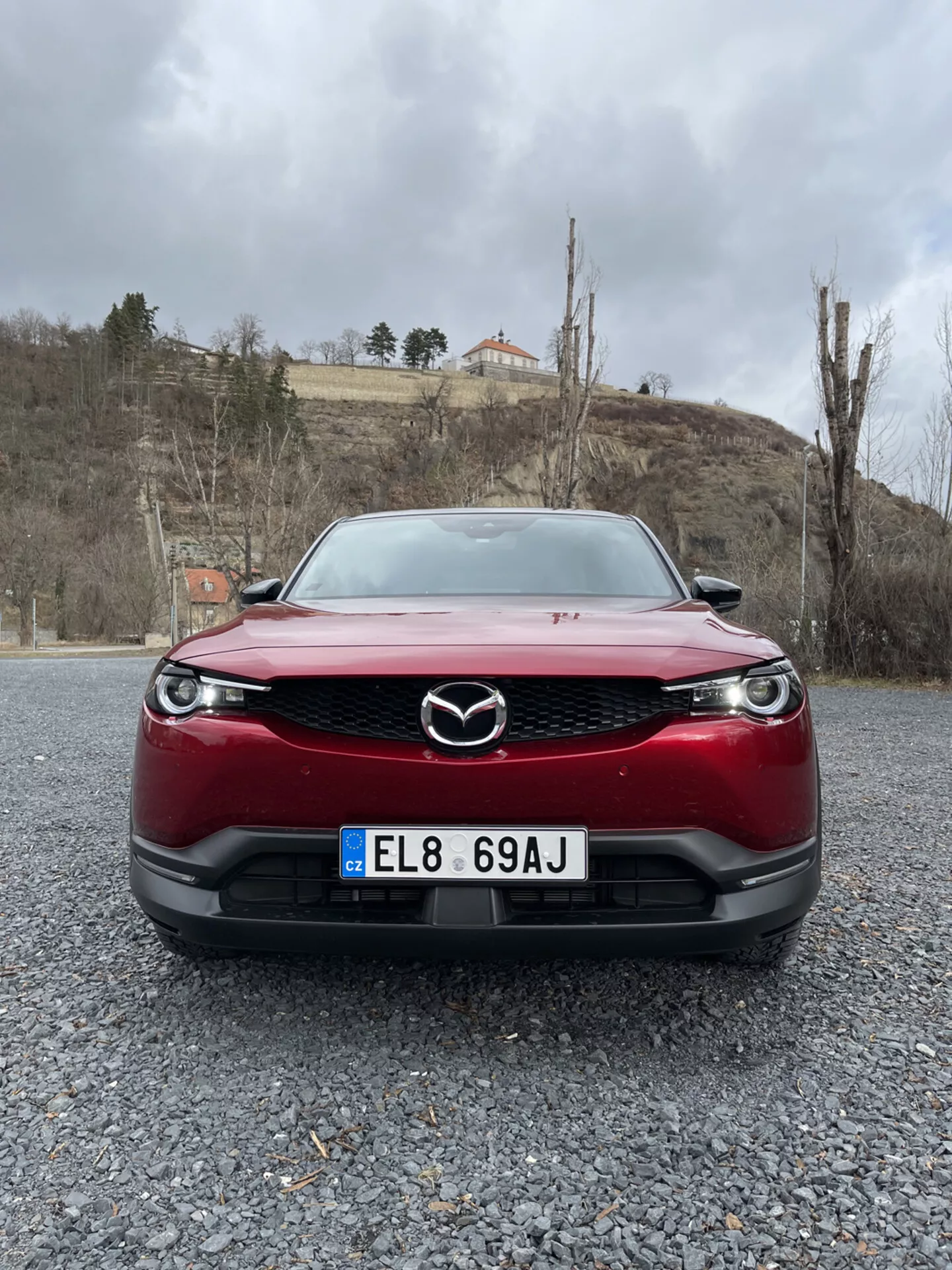 Vista frontale di una Mazda MX-30 del 2020 di colore rosso, con particolare evidenza alla griglia, al paraurti e ai fari LED su una superficie di ghiaia contro uno sfondo di cielo nuvoloso con vegetazione e un edificio