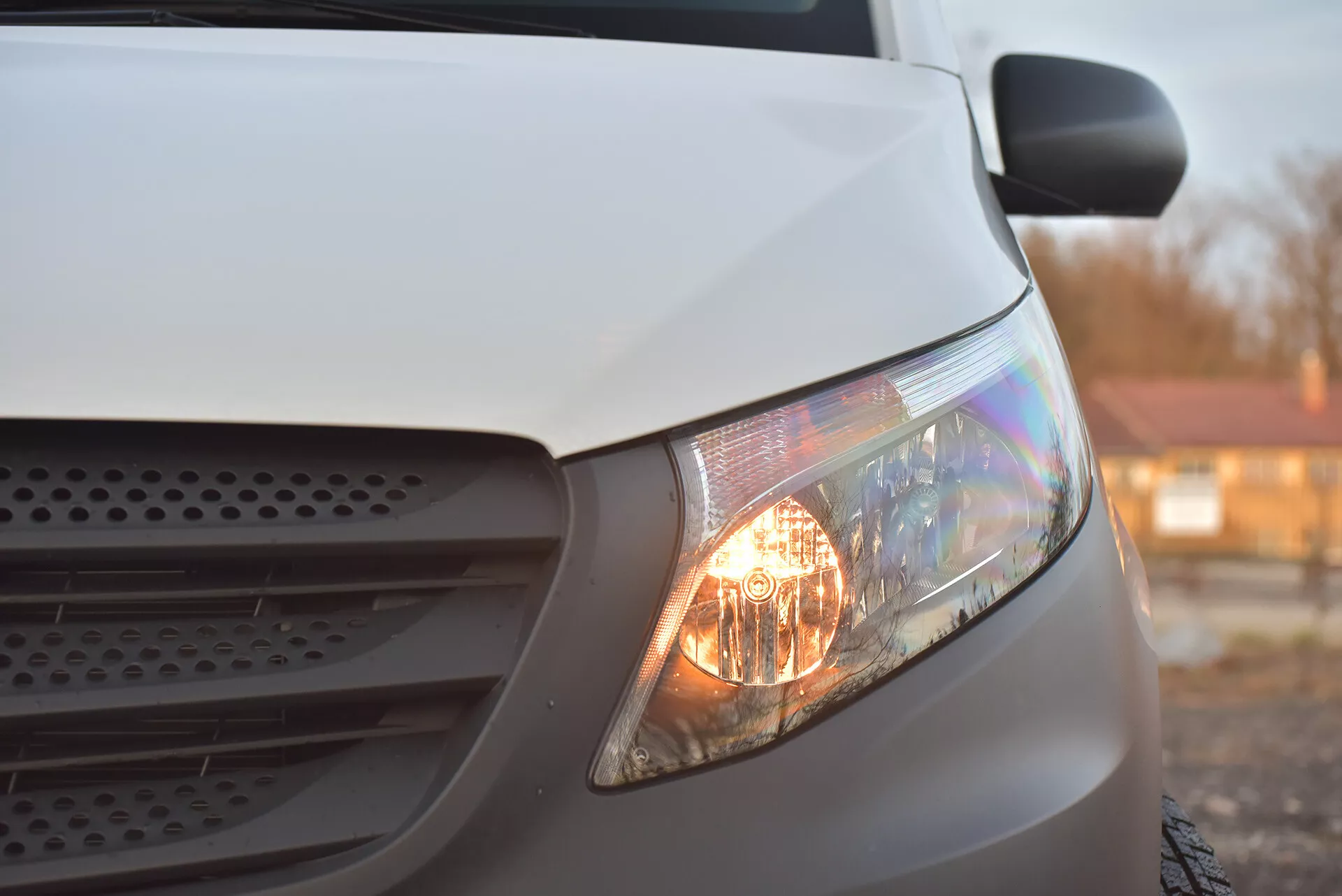 Vista laterale ravvicinata di un Mercedes-Benz Vito Tourer bianco del 2014 focalizzata sul faro illuminato, parte della griglia e sezione del cofano, evidenziando i suoi fari alogeni e design aerodinamico sotto una luce soffusa.