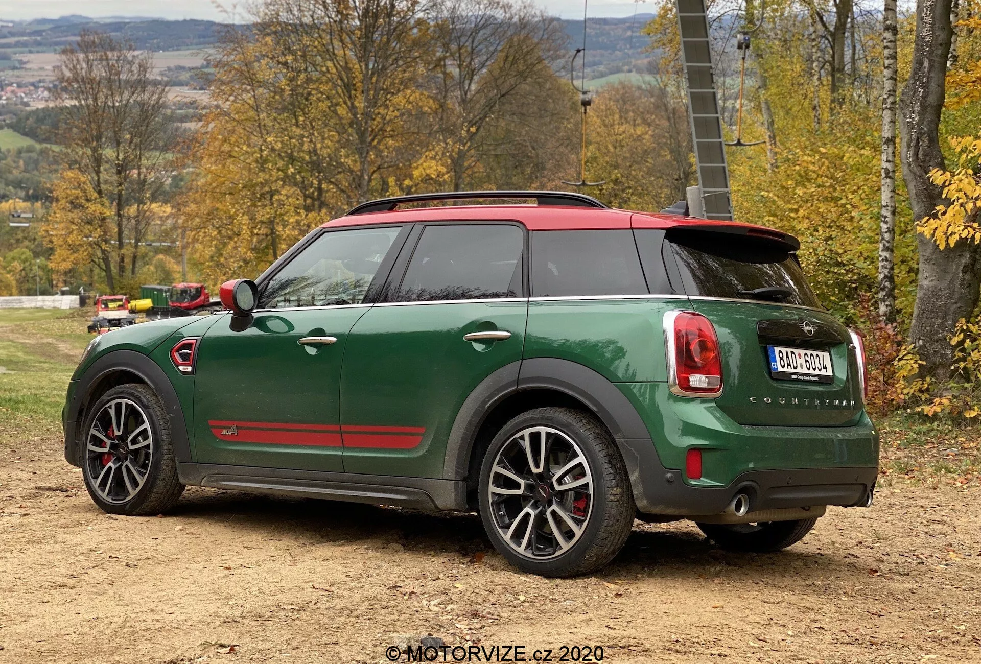 Vista posteriore a tre quarti della Mini Cooper Countryman verde 2020 in edizione sportiva John Cooper Works, con tetto rosso e calotte degli specchietti, doppi tubi di scarico, spoiler posteriore, cerchi in lega a cinque razze con dettagli neri e funzioni tecnologiche avanzate come sensori di parcheggio e antenna a pinna di squalo, posizionata su una superficie di terra in un ambiente rurale autunnale.