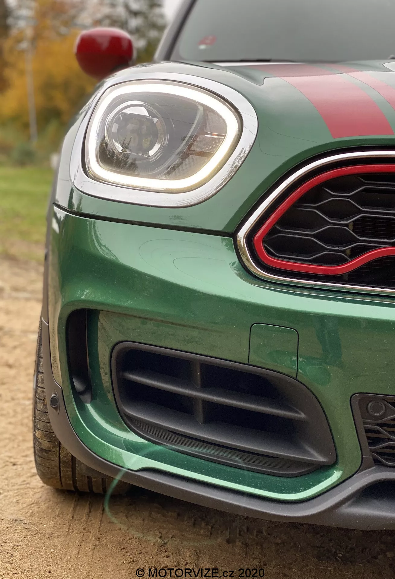 Immagine ravvicinata di una Mini Countryman John Cooper Works 2020 verde vibrante, che si concentra sul quarto anteriore sinistro. Catturata da un'angolazione bassa, l'immagine mette in evidenza la griglia inferiore con design a nido d'ape nero, il paraurti anteriore con caratteristiche aerodinamiche e il faro a LED elegante. La griglia superiore è esagonale con finitura nera lucida e bordo accentuato rosso. L'auto presenta strisce da corsa sul cofano e sul tetto, uno specchietto laterale rosso visibile in alto a sinistra, ed è situata all'aperto con luce naturale e alberi e fogliame sullo sfondo sfocato.