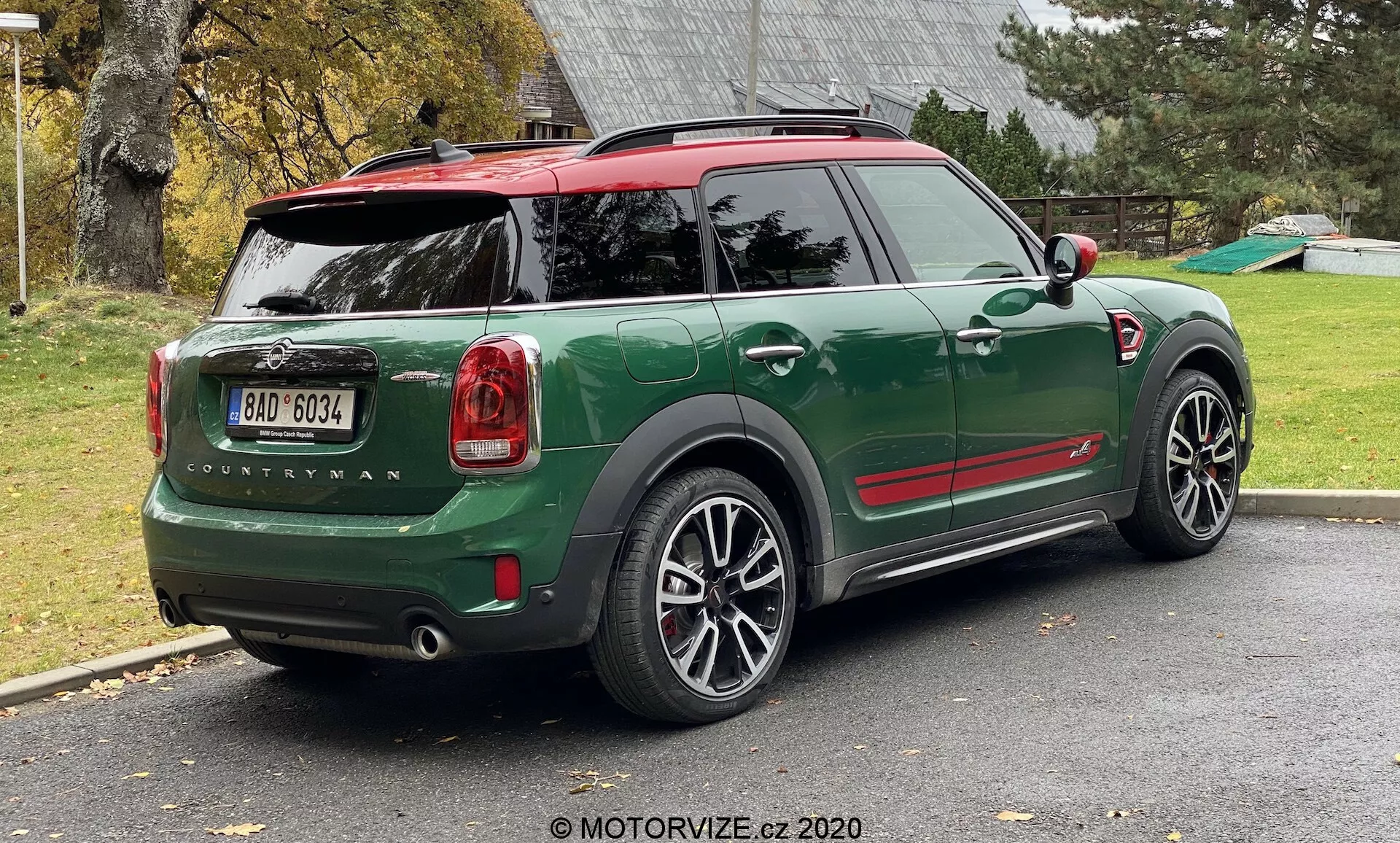 Vista posteriore a tre quarti di un MINI Countryman John Cooper Works 2020 verde parcheggiato su un vialetto residenziale. L'immagine mostra l'intera metà posteriore del lato destro dell'auto, inclusi il portellone posteriore, la ruota posteriore destra, parte del tetto e le porte dei passeggeri. Il design sportivo e compatto del SUV presenta accenti rossi a contrasto sulle calotte degli specchietti laterali e sul tetto, archi delle ruote neri e grandi cerchi in lega a più razze. Sono chiaramente visibili le luci posteriori, il nome del modello 'Countryman' e il logo MINI. L'auto ha un tetto rosso con uno spoiler e barre portatutto, e il lunotto posteriore include uno sbrinatore e un tergicristallo.