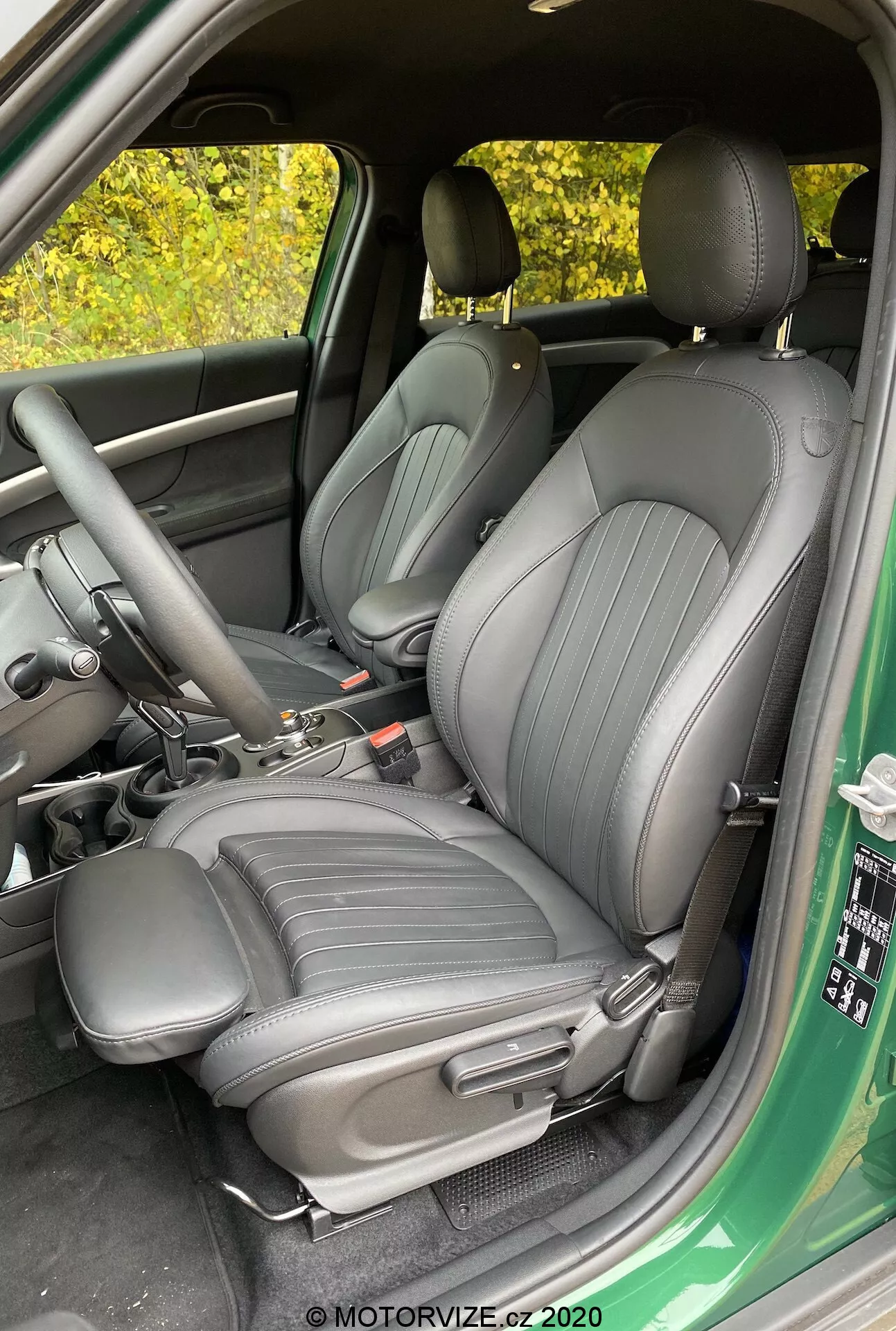 Immagine degli interni della Mini Countryman John Cooper Works 2020 con uno sguardo sui sedili anteriori di guidatore e passeggero, rivestiti in un materiale scuro di alta qualità con linee verticali, vista dalla porta del conducente aperta, che mostra la consolle centrale con leva del cambio e freno a mano, vista parziale del pannello della porta con comandi per i finestrini e gli specchietti.