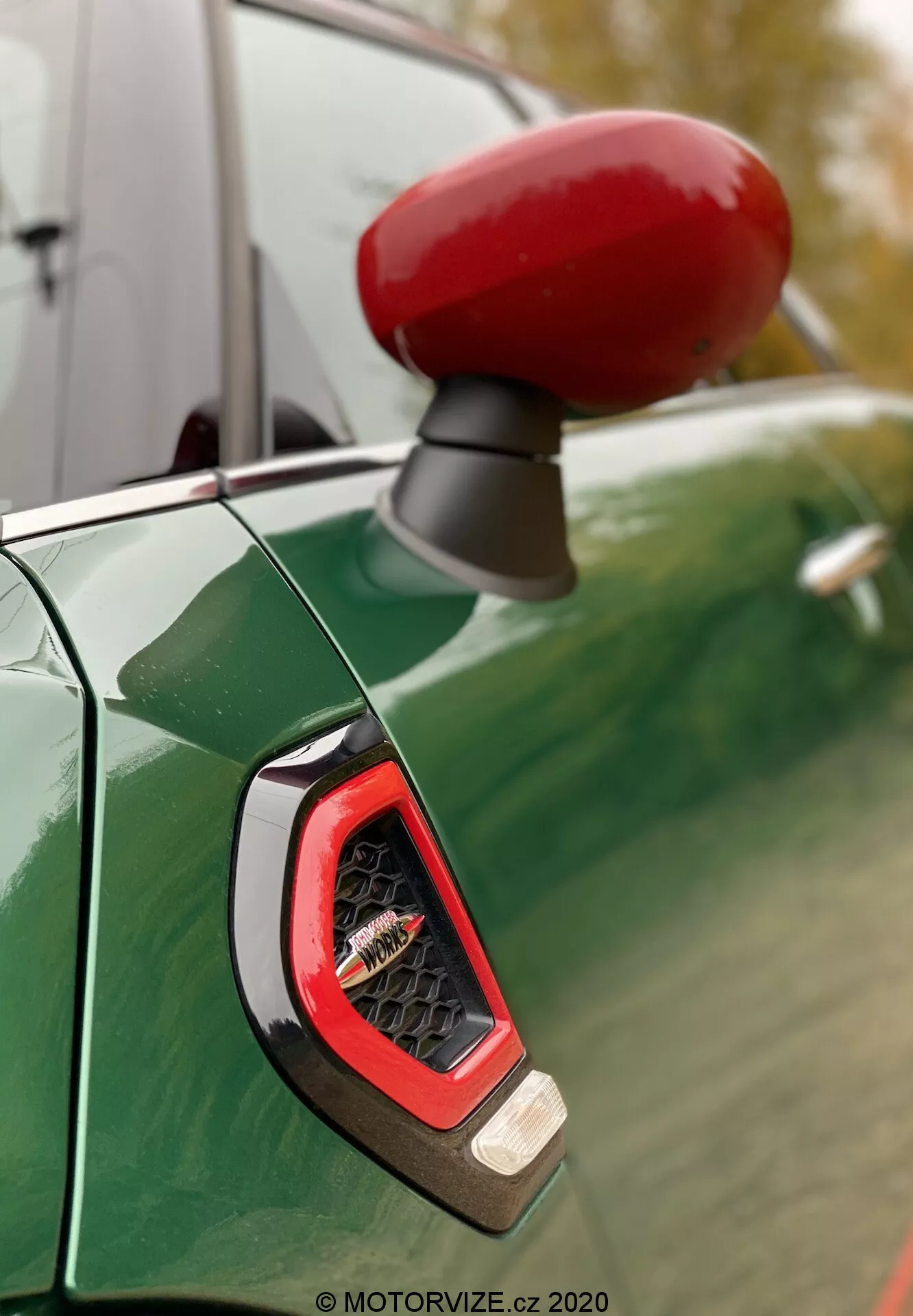 Primo piano dello specchietto laterale di colore rosso vibrante e dell'indicatore laterale sul lato del conducente di una Mini Countryman John Cooper Works 2020, visto contro un pannello della carrozzeria verde metallizzato con riflessi di un cielo nuvoloso e alberi. La messa a fuoco sottolinea l'indicatore angolare con lente trasparente, probabilmente dotato di tecnologia LED, circondato da una finitura sportiva e lucida.