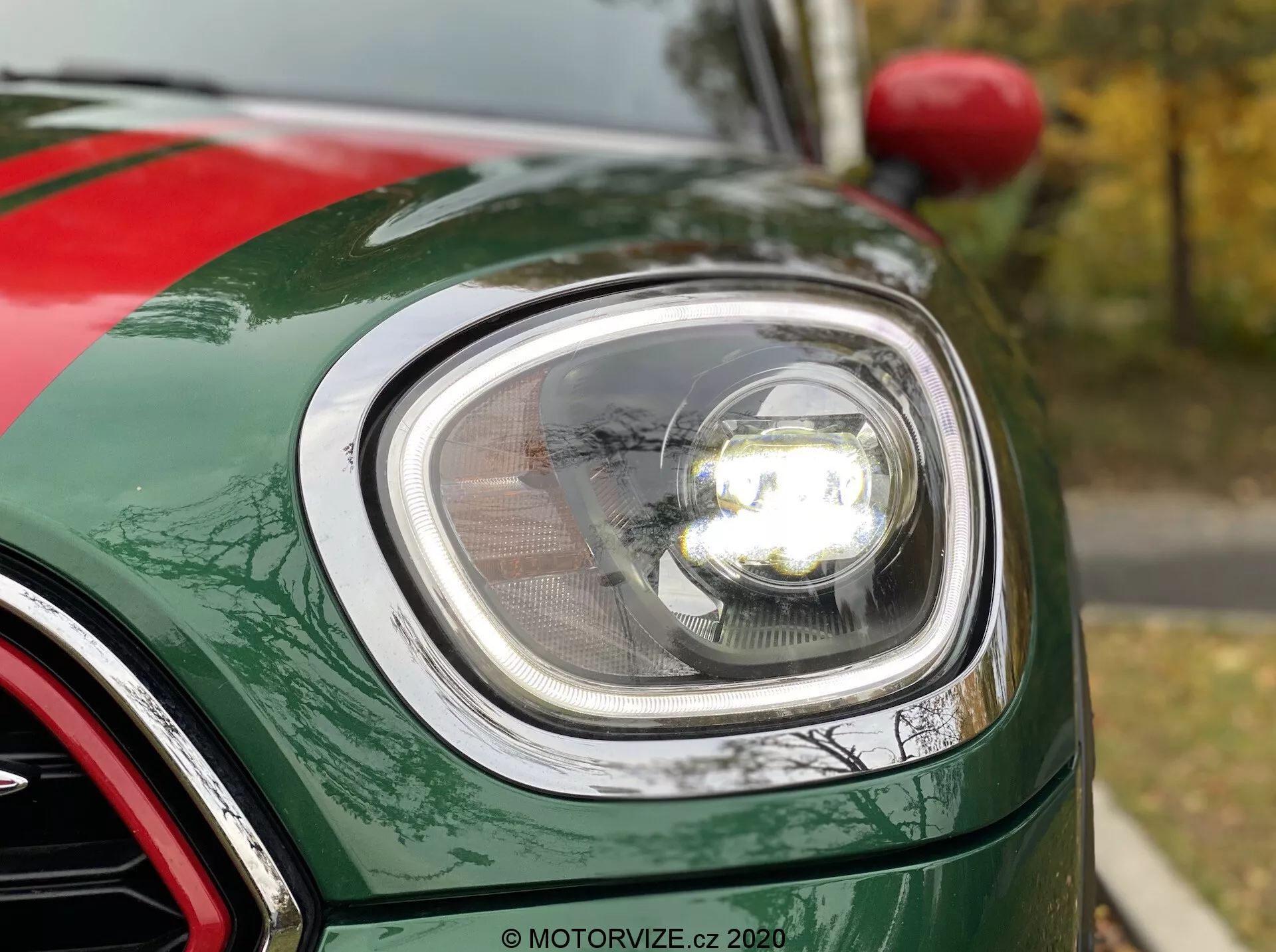 Vista ravvicinata della sezione anteriore sinistra di una 2020 Mini Countryman John Cooper Works, focalizzata sul moderno faro rotondo con tecnologia LED, copertura della lente chiara e faro in stile proiettore illuminato. L'immagine mostra la curvatura del parafango anteriore, il bordo del cofano e una parte della griglia con finitura cromata. L'auto presenta una finitura verniciata verde metallizzato lucido con visibili scaglie metalliche.