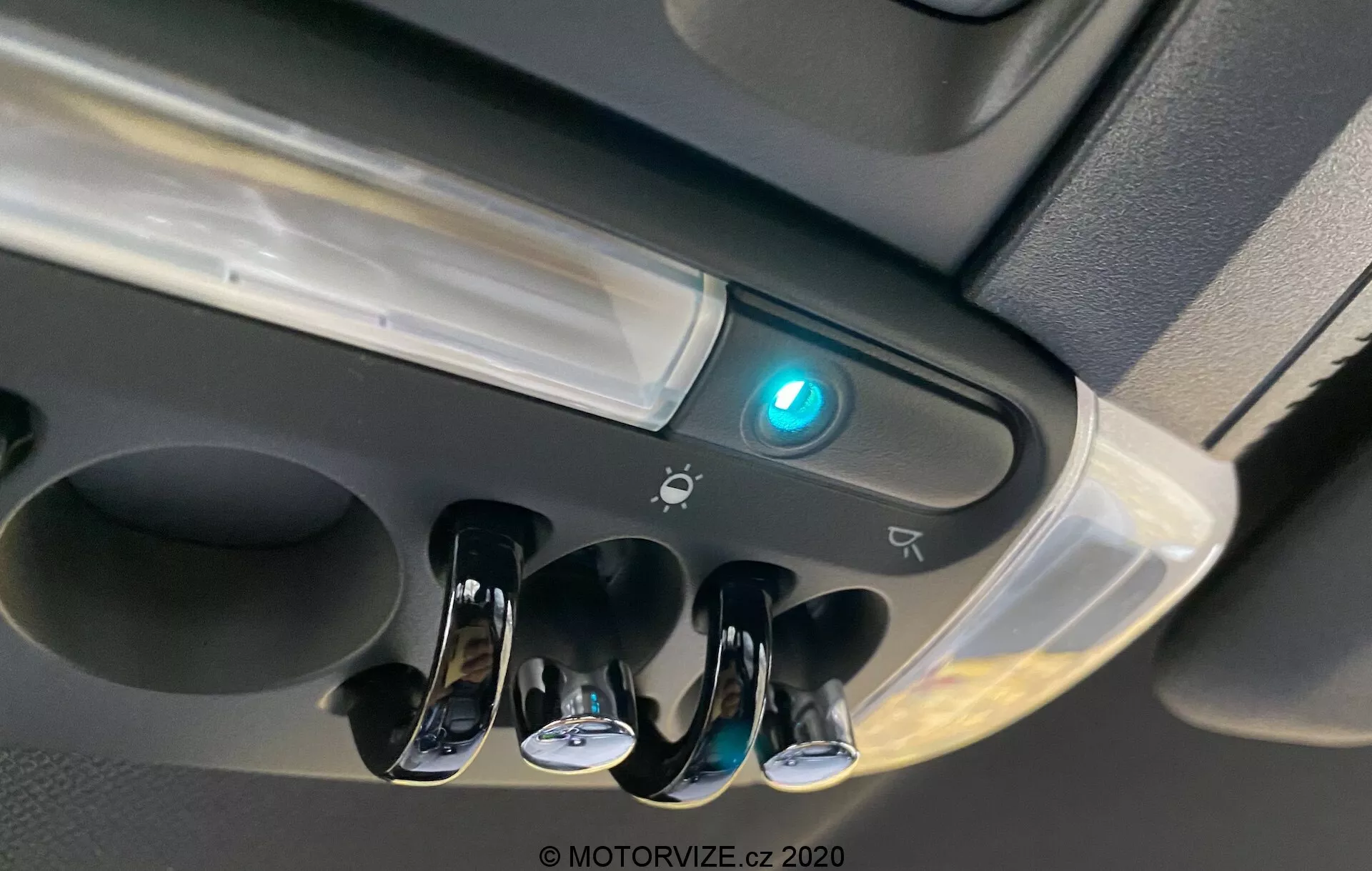 Vista ravvicinata dell'area della console superiore del Mini Countryman 2020 John Cooper Works che mostra le luci interne dell'abitacolo, tre interruttori a levetta con finitura cromata per le luci di lettura individuali, una luce centrale dell'abitacolo, un pulsante illuminato in blu che potrebbe indicare la connettività Bluetooth o una funzione di sicurezza, il bordo di un possibile vano portaoggetti, icone che suggeriscono controlli aggiuntivi o sensori e un design moderno con superfici lucide e opache.