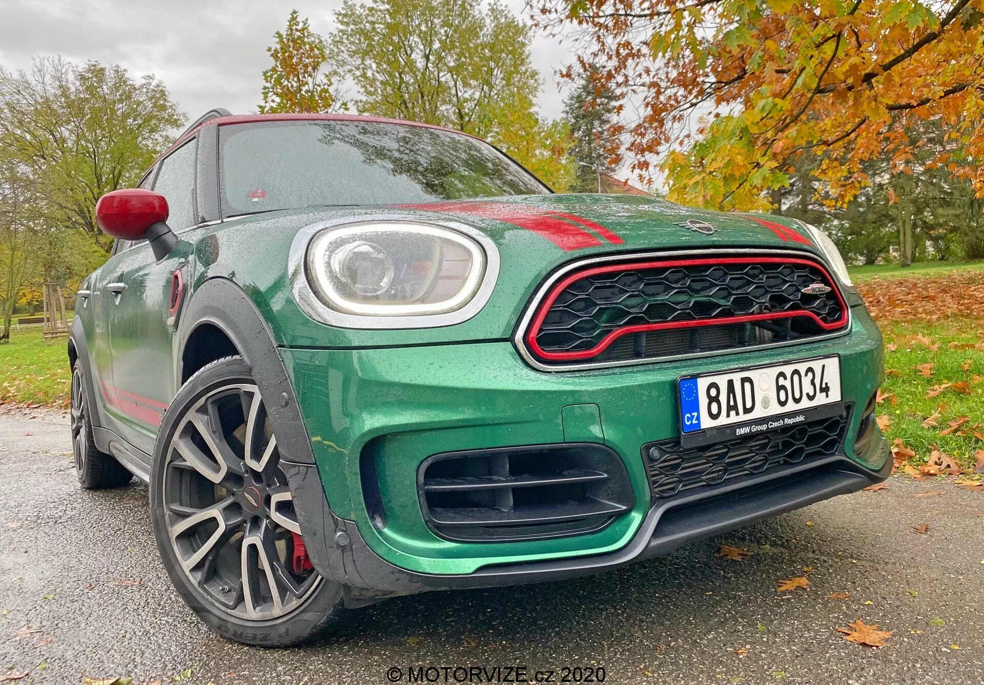 L'immagine presenta la Mini Countryman 2020 in edizione John Cooper Works da un'angolazione frontale bassa, mettendo in risalto il suo design sportivo e moderno. Gli elementi visibili includono la griglia frontale a nido d'ape con accenti rossi, grandi prese d'aria sotto la griglia per migliorare il flusso d'aria, una targa centrale e luci diurne a LED circolari incastonate nei fari principali. Il cofano presenta un leggero rigonfiamento con pieghe aerodinamiche, mentre le linee scolpite nel profilo laterale ne migliorano l'aspetto dinamico. I cerchi in lega a più razze suggeriscono una costruzione leggera per una migliore maneggevolezza e accelerazione, e rivelano pinze e dischi freno ad alte prestazioni. Il corpo è dipinto in un ricco verde metallizzato, accentuato da una calotta dello specchietto laterale rossa e accenti rossi sulla griglia, su uno sfondo autunnale di foglie. Questa prospettiva enfatizza le caratteristiche tecnologiche e di design avanzate dell'auto, rendendola un'automobile di spicco nelle categorie sportive e di lusso.