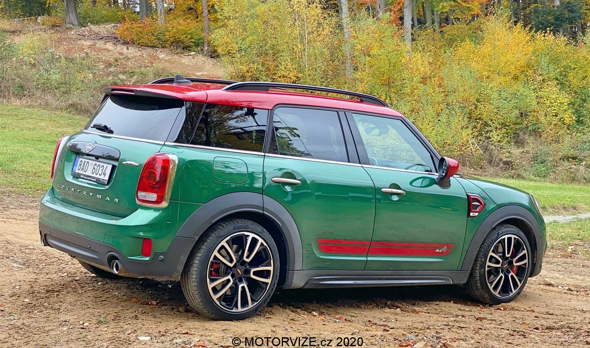 Vista posteriore a tre quarti di una Mini Countryman John Cooper Works 2020 parcheggiata su una superficie non asfaltata con chiazze d'erba. La compatta sportiva presenta uno schema bicolore con carrozzeria verde scuro e tetto e specchietti rossi in contrasto. Il design posteriore mostra un portellone, tergicristallo posteriore, badge prominente e fanali posteriori disposti verticalmente. Il paraurti posteriore include sensori di parcheggio. Dispone anche di cerchi in lega sportivi con design a più razze che mostrano le pinze dei freni, suggerendo una versione ad alte prestazioni. Lo sfondo naturale con alberi autunnali suggerisce la stagione.