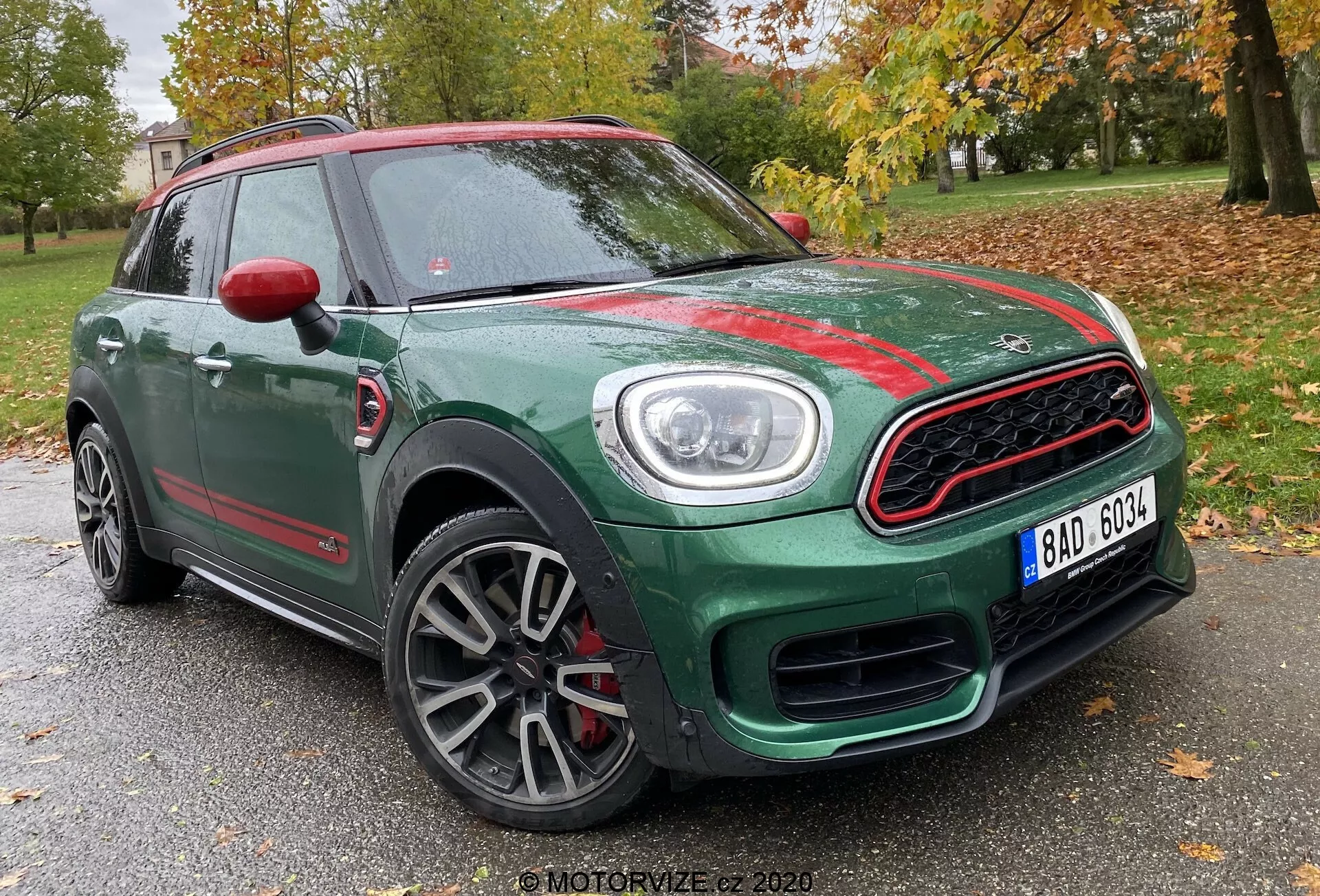 Vista tre quarti anteriore di una Mini Countryman John Cooper Works del 2020, che mostra la sua carrozzeria SUV compatta verde con accenti rossi a contrasto. Catturata da un'angolazione bassa, l'immagine mette in risalto la griglia anteriore sportiva con bordo cromato e design a nido d'ape, fari circolari prominenti con luci diurne a LED, cerchi in lega multirazza con finitura grigia e calotte degli specchietti laterali rosse. La foto è ambientata con uno sfondo di alberi e una struttura tenue, suggerendo una posizione in un parco o area residenziale.