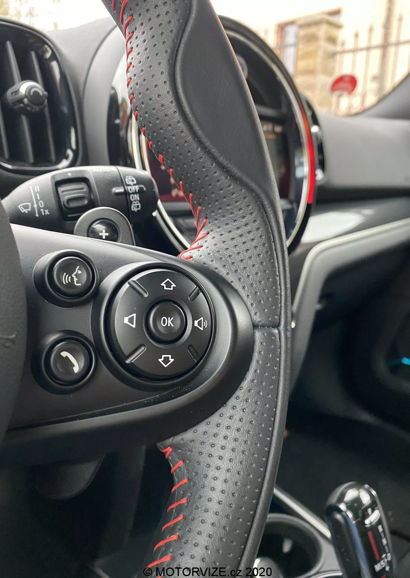 Vista interna di una Mini Countryman John Cooper Works 2020 focalizzata sul volante rivestito in pelle con cuciture rosse, pulsanti di controllo per audio e chiamate e quadro strumenti parzialmente visibile con accenti rossi che indicano un modello sportivo. Mostra l'area di controllo del conducente, il cambio automatico e i controlli dei finestrini elettrici, evidenziando convenienza e funzionalità avanzate.