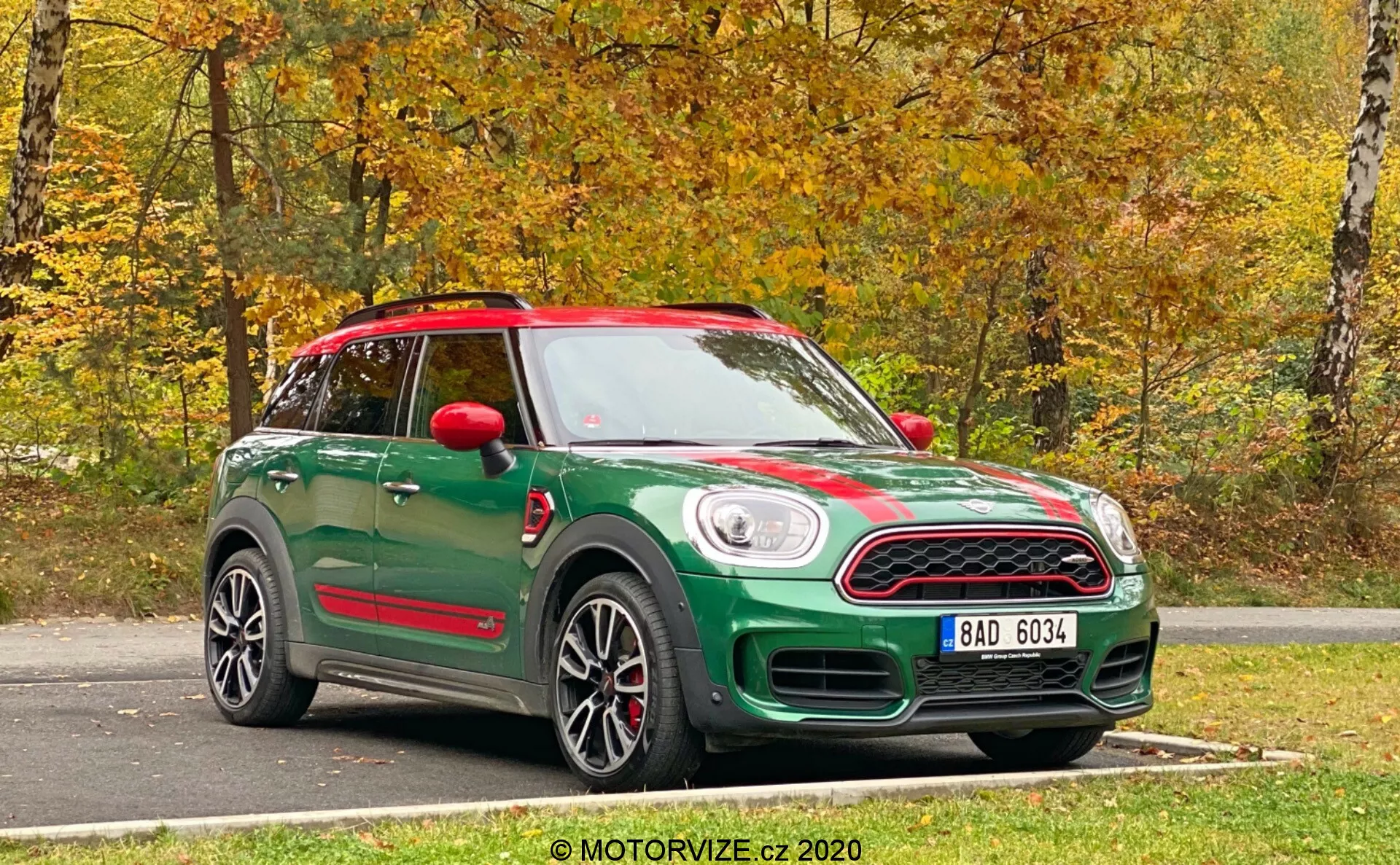Un Mini Countryman John Cooper Works 2020 verde catturato da una vista a tre quarti anteriore. La fotografia mostra sia il frontale che il lato dell'auto, evidenziando le sue caratteristiche di design. La parte anteriore ha una griglia distintiva con una targa sotto di essa e fari rotondi classici. Strisce sportive corrono lungo il cofano, mentre il profilo laterale mostra cerchi in lega, specchietti laterali e la linea del tetto. Gli specchietti laterali rossi e il tetto abbinato contribuiscono a uno schema di colori bicolore. Sotto le porte, minigonne laterali coordinate suggeriscono un pacchetto di finiture aggiornato o sportivo. Il veicolo ha un tetto panoramico ed è parcheggiato su una superficie di asfalto con uno sfondo naturale autunnale. La foto nitida e luminosa è stata scattata alla luce naturale.