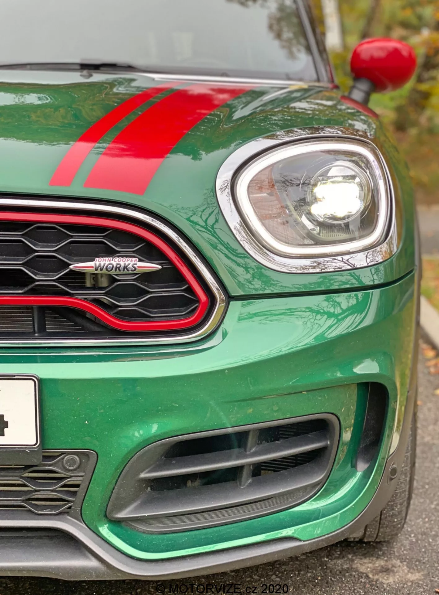 Vista ravvicinata della parte anteriore sinistra di una Mini Countryman John Cooper Works 2020. L'immagine, scattata da leggermente sotto il livello dei fari, mette in evidenza il cofano verde metallizzato con una striscia da corsa rossa, il faro sinistro circolare incorniciato in cromo e una parte della griglia a nido d'ape con contorno nero lucido. Sono visibili il badge 'John Cooper Works', l'indicatore sinistro sull'arco della ruota e le grandi prese d'aria del paraurti anteriore progettate per il raffreddamento del motore e le prestazioni aerodinamiche. L'auto appare pulita e ben curata, sottolineando la sua tecnologia di fari a proiettore o LED moderni e il design sportivo orientato alle prestazioni.