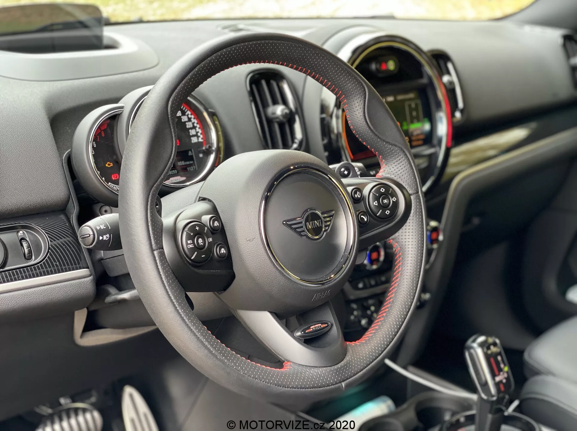 Vista interna del Mini Countryman John Cooper Works 2020 dalla prospettiva del conducente, con un volante in pelle a fondo piatto con cuciture visibili e controlli multifunzione, palette al volante, doppi quadranti circolari degli strumenti che fiancheggiano un display digitale centrale, e un cruscotto con vari pulsanti e interruttori nella console centrale.