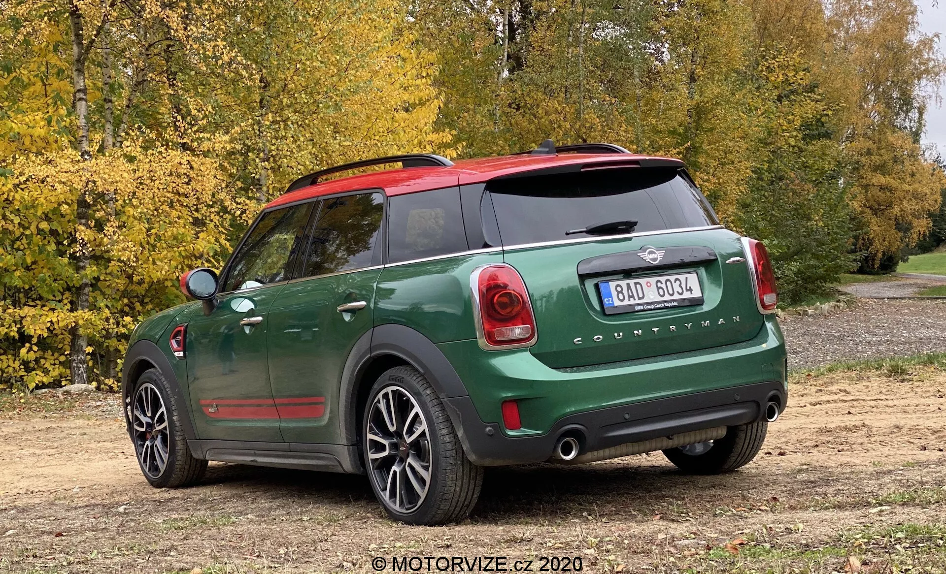 Vista posteriore di tre quarti di una Mini Cooper Countryman John Cooper Works 2020 con vernice bicolore verde scuro e nera, cappucci degli specchietti rossi, emblema Mini visibile e scritta 'Countryman', fanali posteriori dal design unico, paraurti posteriore prominente con sensori di parcheggio, finestrino posteriore con sbrinatore, doppi tubi di scarico, tergicristallo posteriore, cerchi in lega a cinque razze, parcheggiata su un sentiero sterrato con alberi e foglie autunnali in sottofondo, catturata da un angolo leggermente alto.