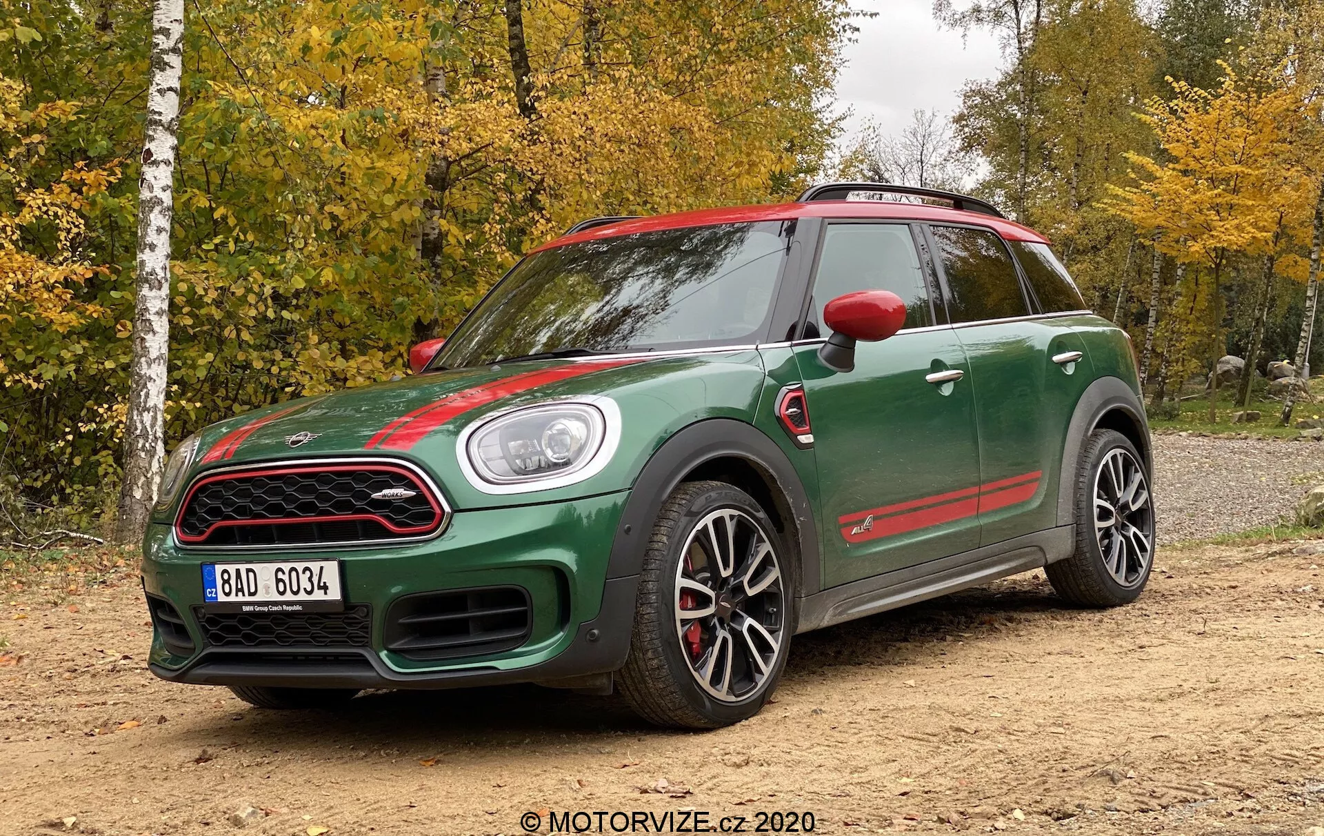 Vista angolare anteriore di tre quarti di un Mini Countryman John Cooper Works 2020, con design sportivo con carrozzeria bicolore verde scuro e accenti rossi sugli specchietti laterali, la griglia anteriore e le minigonne laterali. Caratteristiche includono una presa d'aria sul cofano distintiva, fari rotondi, griglia frontale a nido d'ape, grandi ruote in lega sportive con design a più raggi, pinze freno rosse, minigonne laterali pronunciate e una postura aggressiva. L'auto è posizionata su una superficie non asfaltata con uno sfondo forestale, sotto una luce naturale in una giornata nuvolosa.