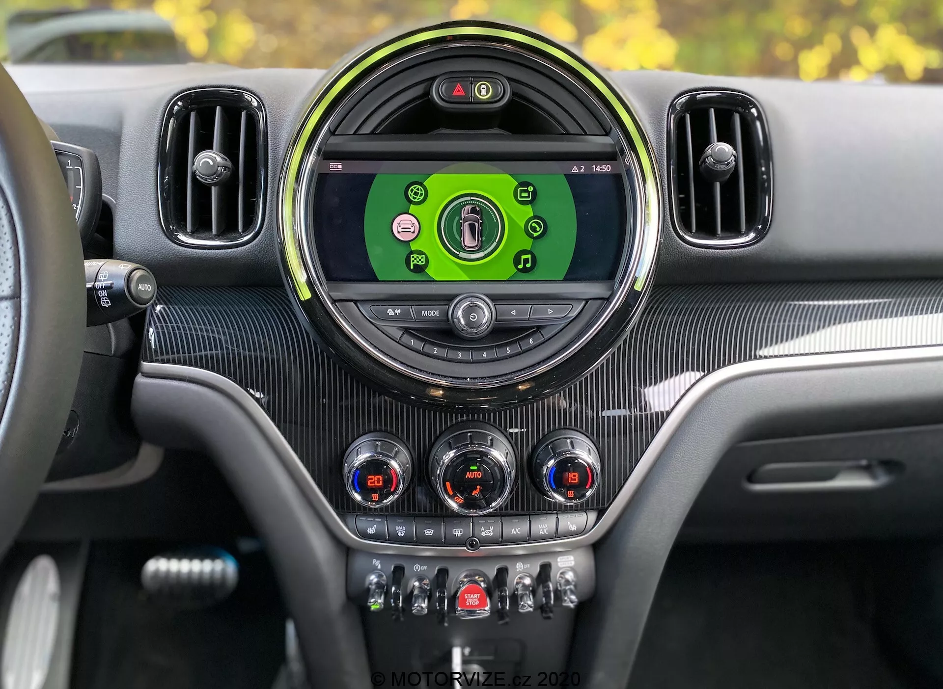 Cruscotto interno del Mini Countryman John Cooper Works 2020 dalla prospettiva del conducente. Consolle centrale con grande display circolare per l'infotainment che mostra icone colorate per media, connettività telefonica, navigazione e impostazioni del veicolo. Incorniciato da una cornice circolare elegante e affiancato da ulteriori prese d'aria. Sotto il display ci sono quadranti analogici con accenti cromati per il controllo del clima e interruttori per funzioni come riscaldamento dei sedili, sbrinamento, ricircolo dell'aria e modalità di guida. Texture tipo fibra di carbonio e accenti metallici danno un design moderno e lussuoso con quadro strumenti digitale visibile che indica che il veicolo è acceso.