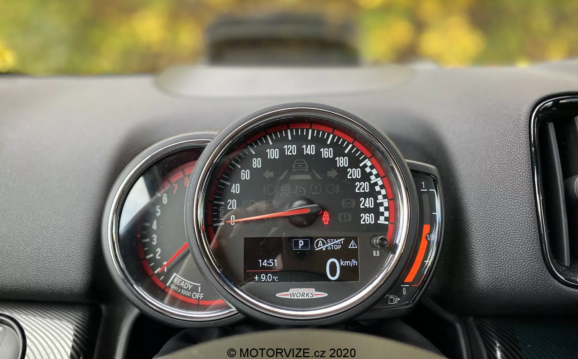 Primo piano dettagliato del cruscotto del Mini Countryman John Cooper Works 2020. Il focus è su un tachimetro circolare che va da 0 a 260 km/h, con la lancetta a 0 km/h, indicando che l'auto è ferma. Un display digitale in basso mostra l'auto in modalità 'P', una temperatura esterna di 9,0°C, un chilometraggio di 1451 km e l'icona del bagagliaio aperto. Le parole 'Auto Start/Stop' suggeriscono un sistema di risparmio carburante. Il tachimetro è incorniciato da un anello cromato e ha una zona rossa che inizia subito dopo i 220 km/h. A sinistra, c'è una vista parziale di un altro indicatore, probabilmente un contagiri. Il cruscotto ha una superficie testurizzata e una piccola bocchetta d'aria è parzialmente visibile a destra del tachimetro.