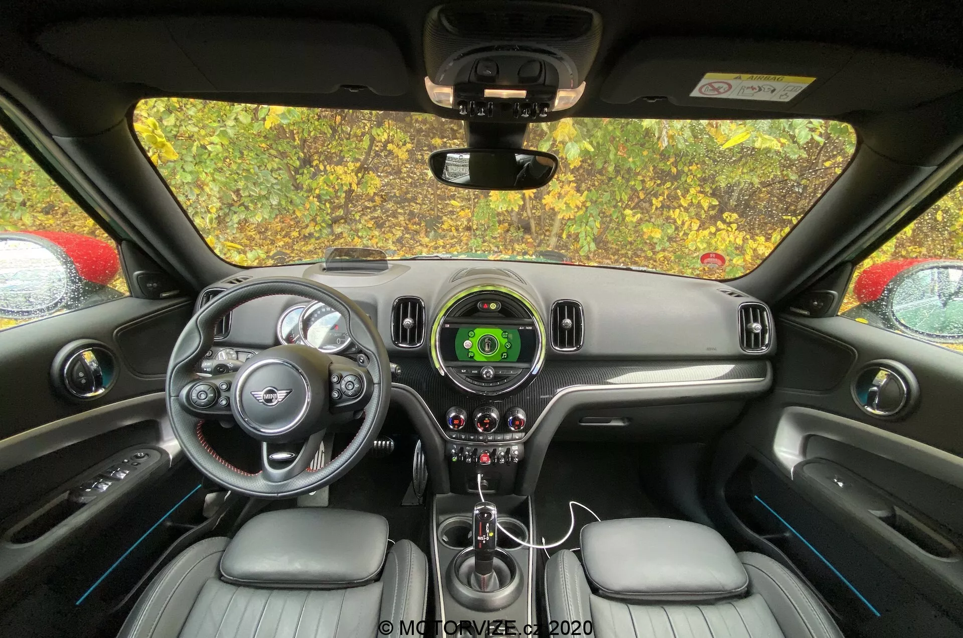 Interni del Mini Countryman 2020 John Cooper Works dalla prospettiva del sedile posteriore, focalizzandosi sul cruscotto e sull'area di controllo del conducente. Sono visibili il volante con accenti argentati e logo del marchio, tachimetro centrale con display digitale, design elegante del cruscotto, leva del cambio, controlli del clima, sedili rivestiti in pelle e specchio retrovisore.