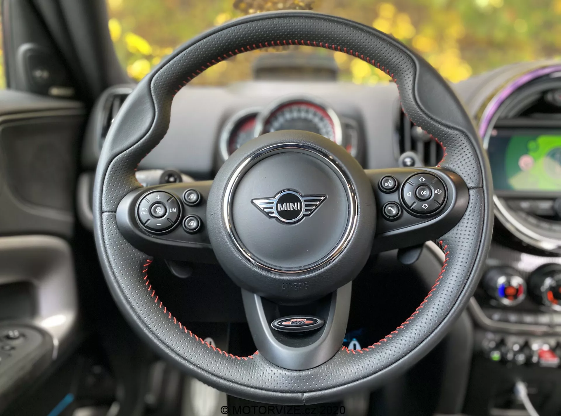 Vista interna di una Mini Countryman John Cooper Works 2020 dal sedile del guidatore, focalizzata sul volante con il logo Mini al centro. Il volante a tre razze, rivestito in pelle con cuciture rosse, dispone di comandi multifunzionali per audio, telefono, controllo vocale e controllo della velocità. Sono visibili le palette del cambio, che indicano una trasmissione automatica con selezione manuale delle marce. Sullo sfondo si distingue il cruscotto con elementi di design circolari, un display centrale che mostra una mappa, bocchette dell'aria condizionata e accenti colorati.