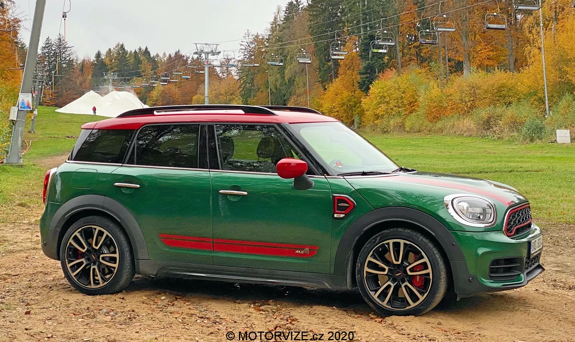 Profilo laterale di una Mini Cooper Countryman 2020 John Cooper Works verde posizionata su terreno sabbioso con alberi autunnali e infrastruttura di una seggiovia sullo sfondo. L'auto presenta un tetto bianco, copri specchietti rossi, ruote multiraggio nere e argento aggressive con pinze freno rosse e minigonne laterali sportive rosse.