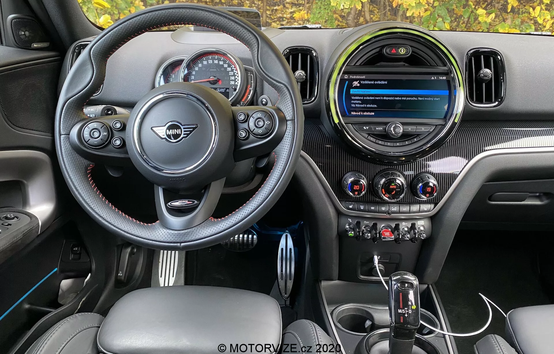 Vista dell'interno del Mini Countryman John Cooper Works 2020, evidenziando la plancia del conducente con volante sportivo a fondo piatto, pulsanti multifunzionali, cuciture a contrasto e stemma centrale del produttore. La plancia include un moderno schermo infotainment circolare, tre prese d'aria metalliche e pulsanti di controllo del clima ben integrati. La console centrale presenta una leva del cambio automatico con cuffia in pelle e una manopola di controllo infotainment, il tutto completato da texture in fibra di carbonio, pelle e accenti metallici per un design premium e orientato alle prestazioni.