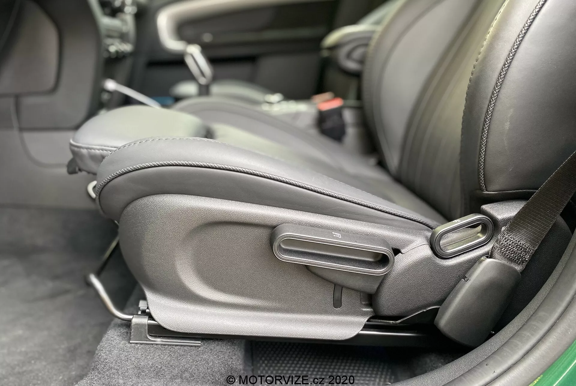 Vista ravvicinata dell'interno della Mini Countryman John Cooper Works 2020, evidenziando la parte inferiore laterale del sedile del conducente con dettagliati meccanismi di regolazione del sedile. Il sedile è rivestito in pelle scura o similpelle con un supporto laterale prominente, evidenziando comfort e qualità. Sono visibili i comandi di regolazione manuale, tra cui una maniglia a barra e una manopola rotonda, suggerendo un design ergonomico e regolabile dall’utente. È presente anche un ricevitore della cintura di sicurezza, indicando attenzione alla sicurezza dei passeggeri.