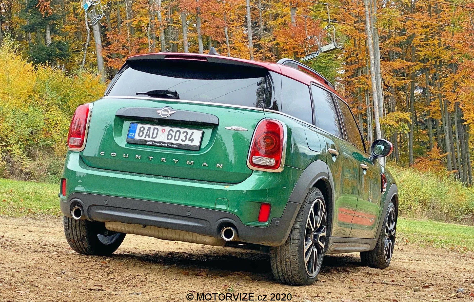 Immagine di una Mini Countryman 2020 modello John Cooper Works verde parcheggiata all'aperto in un contesto autunnale, presa da un angolo posteriore tre quarti. Il lato posteriore destro dell'auto è rivolto verso la fotocamera e mostra le luci posteriori, il parabrezza posteriore, il badge 'COUNTRYMAN', il doppio sistema di scarico, le ruote a cinque razze, il tetto nero a contrasto e gli specchietti laterali. L'immagine include anche una targa posteriore, un tergicristallo posteriore e un'antenna per la connettività.