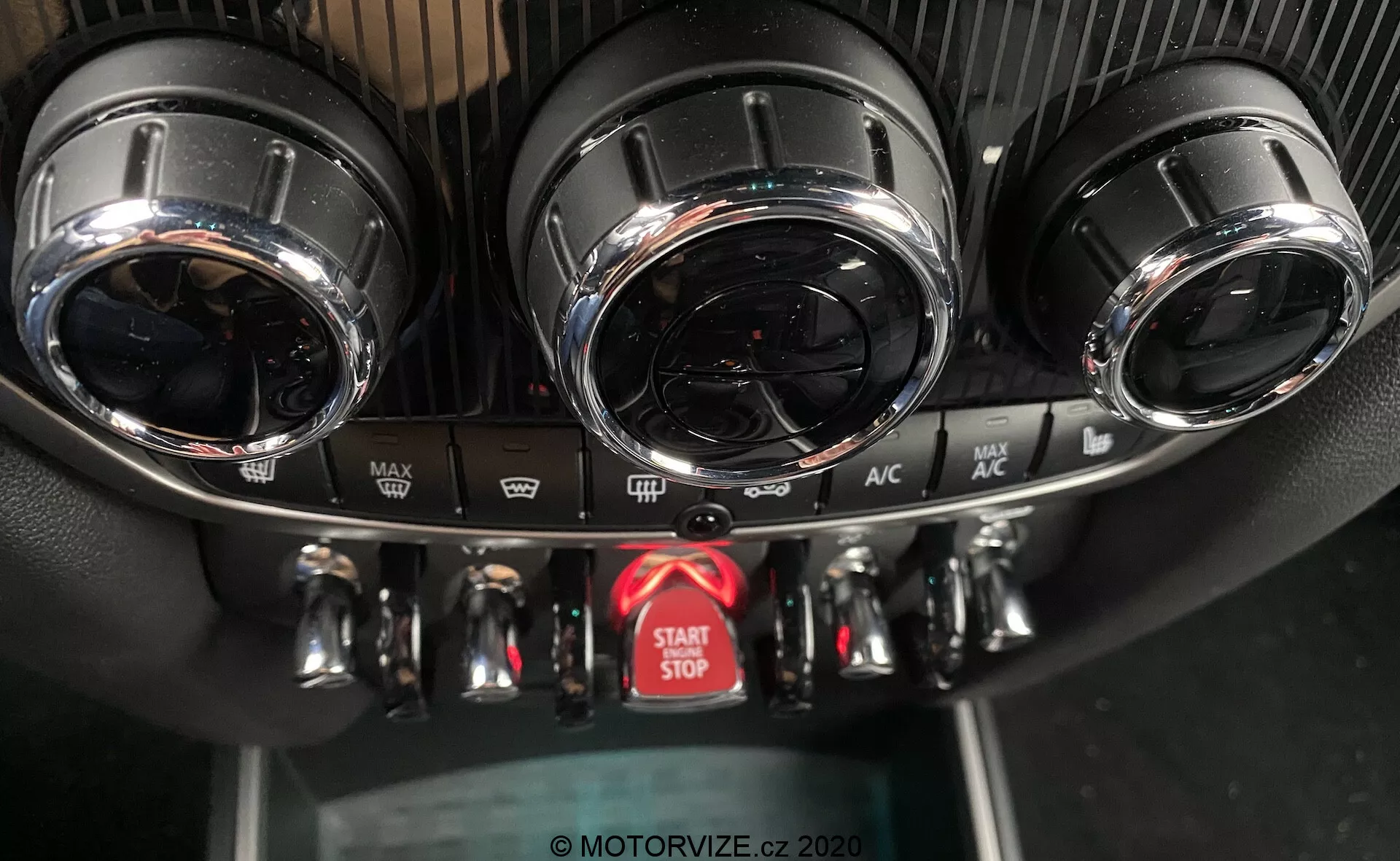 Questa immagine cattura una vista ravvicinata della console centrale del cruscotto in una Mini Countryman John Cooper Works del 2020, evidenziando il sistema di controllo del clima e il pulsante di avvio/arresto del motore. La console mostra due prese d'aria circolari con finiture metalliche nella parte superiore, un pannello di controllo del clima dettagliato con tre manopole in metallo per la temperatura, la velocità della ventola e la direzione del flusso d'aria, e vari pulsanti per funzioni come sbrinamento e ricircolo dell'aria. Sotto i controlli si trova un pulsante rosso ben visibile per l'avvio e l'arresto del motore. Le texture del pannello includono un design sportivo simile alla fibra di carbonio intorno alle prese d'aria e plastica nera standard per il pannello inferiore. L'angolazione della foto, dal davanti e leggermente dall'alto, enfatizza il design di alta gamma e le caratteristiche avanzate.