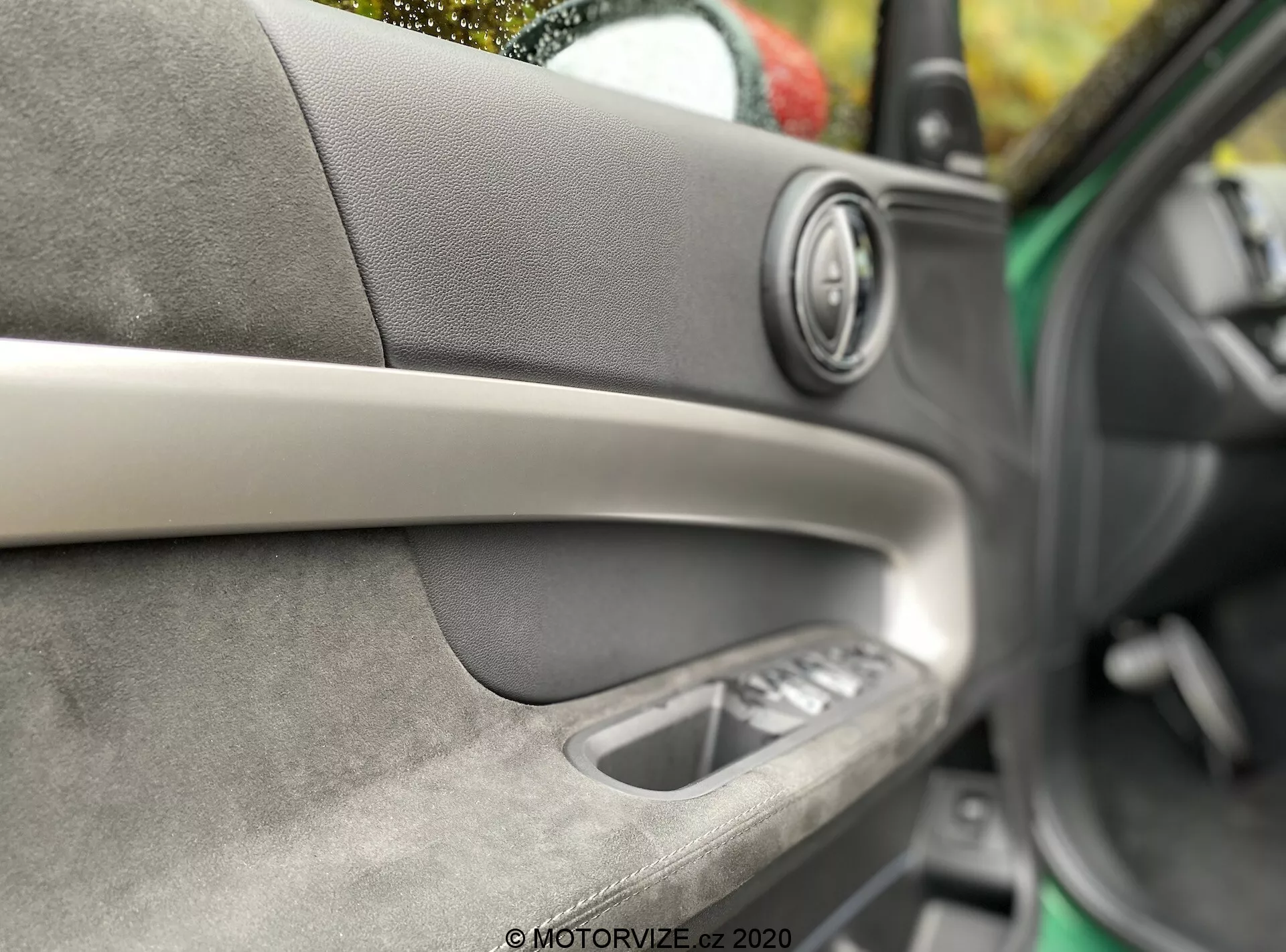 Dettaglio dell'interno della porta di una Mini Countryman John Cooper Works 2020. Messa a fuoco sull'altoparlante audio premium con anello metallico, striscia decorativa diagonale, materiale morbido al tatto con cuciture a vista, maniglia interna della porta e controlli dei finestrini elettrici. Profondità di campo bassa che evidenzia la maestria artigianale e il comfort degli interni.