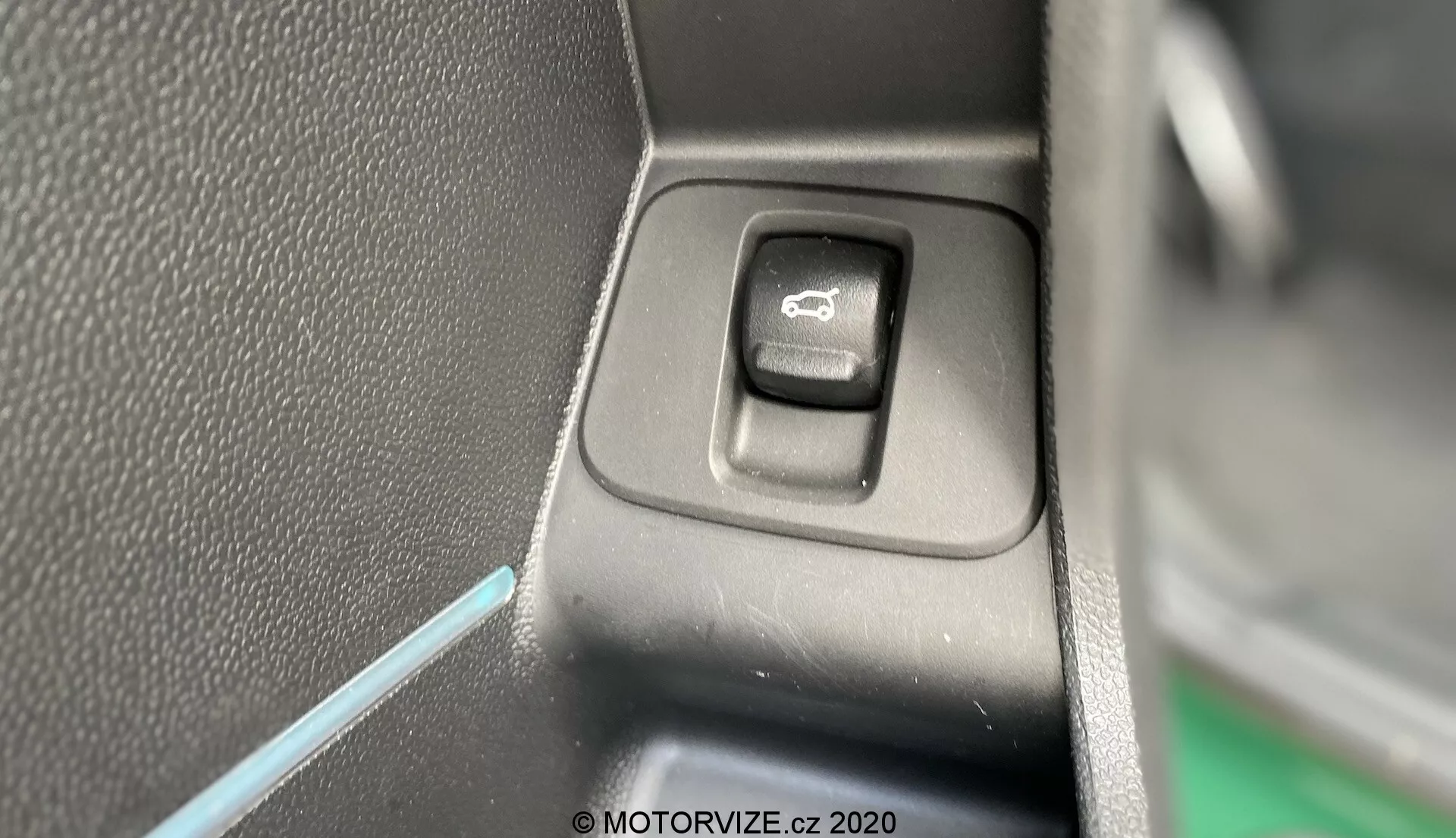 Vista ravvicinata dell'interruttore di controllo per azionare gli specchi laterali nell'interno del Mini Countryman 2020 John Cooper Works. L'interruttore rettangolare con angoli arrotondati, situato su un pannello piatto, è evidenziato con un simbolo a specchio. Un lato dell'interruttore è in ombra, indicando una fonte di luce dal lato opposto. Lo sfondo presenta plastica nera testurizzata e una sottile linea di luce ambientale blu, contribuendo all'estetica degli interni del veicolo.