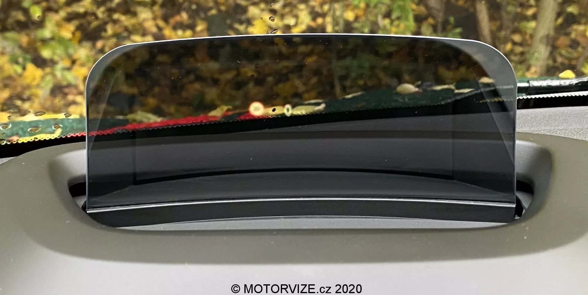 Immagine che mostra l'head-up display (HUD) della Mini Countryman 2020, proiettato su uno schermo trasparente sopra il cruscotto dalla prospettiva del conducente. Il pannello HUD, chiaro e simile al vetro, è perfettamente integrato nel design degli interni, arrotondato e leggermente inclinato per una visibilità ottimale. Lo sfondo fuori dal parabrezza è sfocato, mettendo in evidenza la concentrazione dell'HUD nel presentare informazioni cruciali per la guida.