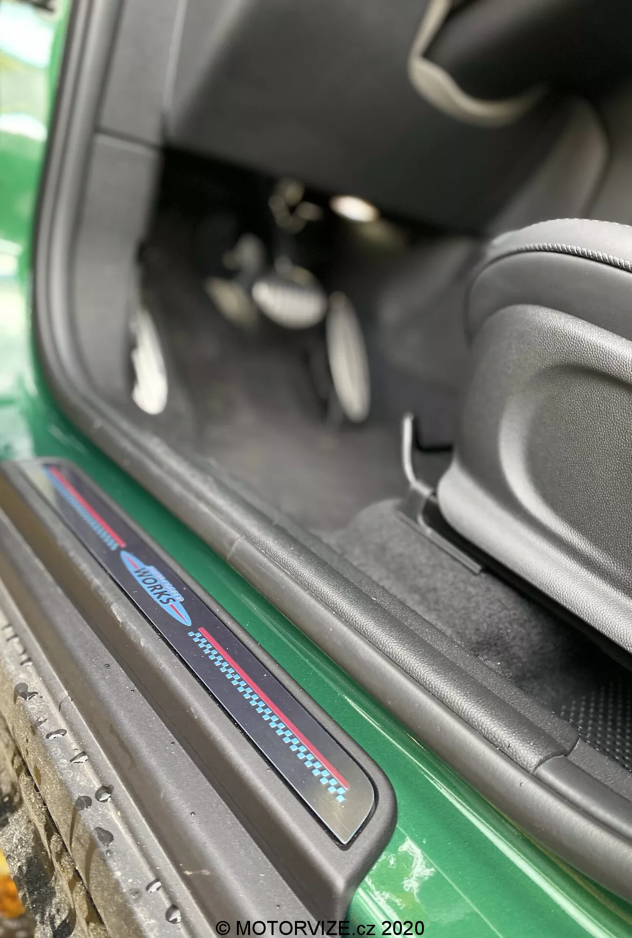 Vista ravvicinata dell'area della soglia della porta della Mini Countryman John Cooper Works 2020 da un angolo obliquo focalizzata sulla soglia del lato passeggero. L'immagine, catturata dall'esterno del veicolo, evidenzia la parte inferiore dell'interno dell'auto, inclusa la piastra della soglia con dettagli blu e rossi, tappeti neri, il supporto laterale sportivo del sedile nero, una vista parziale dei pedali e un'area della soglia ben mantenuta. Gli elementi esterni visibili includono una minigonna laterale verde e la carrozzeria, migliorando l'appello estetico. La filigrana 'MOTORVIZE.cz 2020' suggerisce che la foto è stata scattata nel 2020 per una recensione automobilistica.