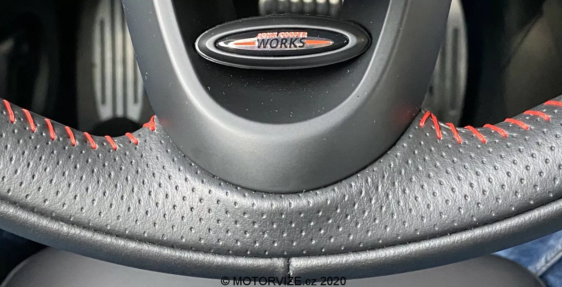 Vista ravvicinata del volante dalla prospettiva del sedile del conducente, con il logo 'John Cooper Works', rivestimento in pelle con trama a fossette per una presa migliorata e cuciture a contrasto rosse indicative delle varianti ad alte prestazioni della MINI Cooper