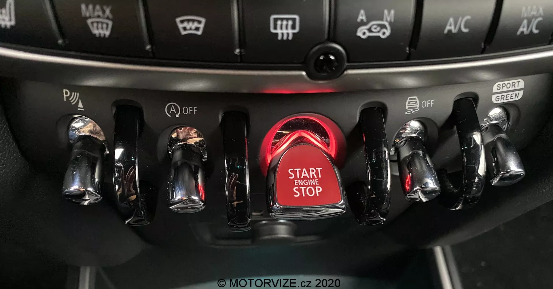 Vista ravvicinata della console centrale del Mini Countryman John Cooper Works 2020 con un focus sull'area di accensione, evidenziando il pulsante di avvio/arresto del motore circondato da un anello rosso luminoso, con interruttori metallici finiti in cromo su entrambi i lati per vari controlli del veicolo, inclusi i modi di guida e le impostazioni del clima.