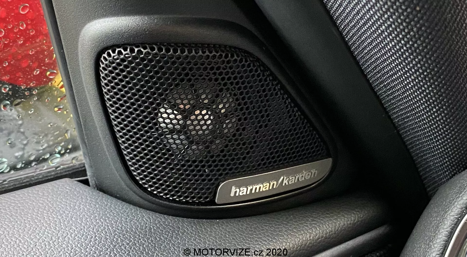 Vista ravvicinata del sistema audio Harman Kardon all'interno del pannello porta di una Mini Countryman 2020 John Cooper Works. La fotografia si concentra sulla griglia del diffusore perforata, progettata per trasparenza acustica e protezione, contro un interno con texture simile alla pelle. Gocce di pioggia sono visibili sul finestrino della macchina, aggiungendo contesto all'immagine. Il sistema audio premium evidenzia caratteristiche tecnologiche chiave e mostra il lusso e la durabilità del design del veicolo.