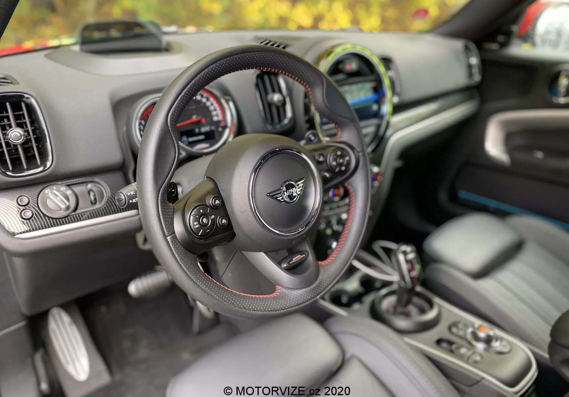 Vista interna del Mini Countryman 2020 dalla prospettiva del conducente, con particolare attenzione al volante multifunzione con cuciture rosse, al quadro strumenti con display analogici e digitali, alle bocchette dell'aria condizionata e al display dell'infotainment. Sulla console centrale sono visibili la leva del cambio, i portabicchieri, i pulsanti di controllo e il portachiavi che indica l'ingresso senza chiave e l'avviamento con pulsante. Il primo piano è focalizzato sul volante e sulla leva del cambio, mostrando l'ergonomia e il comfort progettati per il conducente.