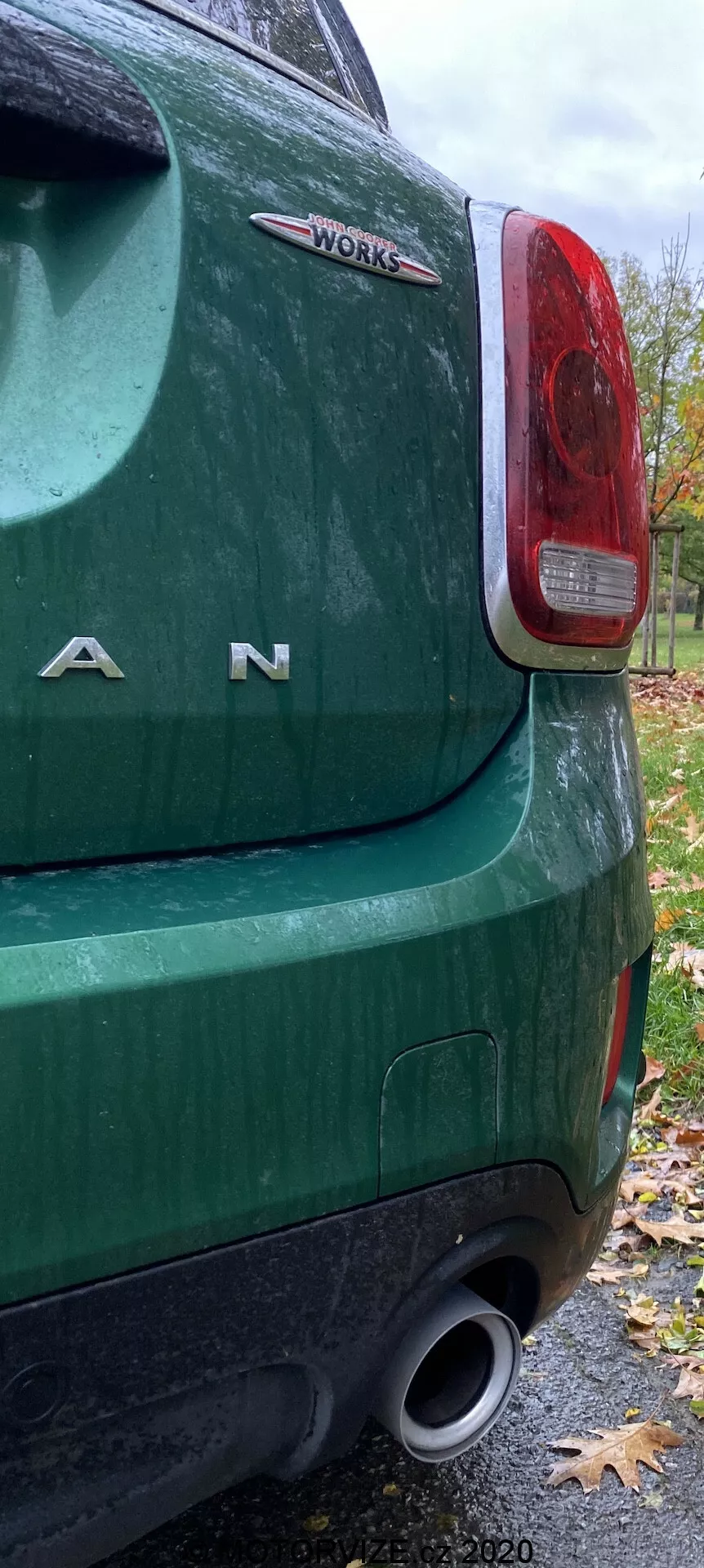 Vista ravvicinata dell'angolo posteriore sinistro di una Mini Countryman John Cooper Works 2020 verde, evidenziando il fanale posteriore, le lettere cromate 'MINI', il badge 'WORKS', il paraurti posteriore e lo scarico, indicando caratteristiche ad alte prestazioni. L'immagine è stata scattata in una giornata autunnale nuvolosa, mostrando superfici bagnate e foglie cadute.