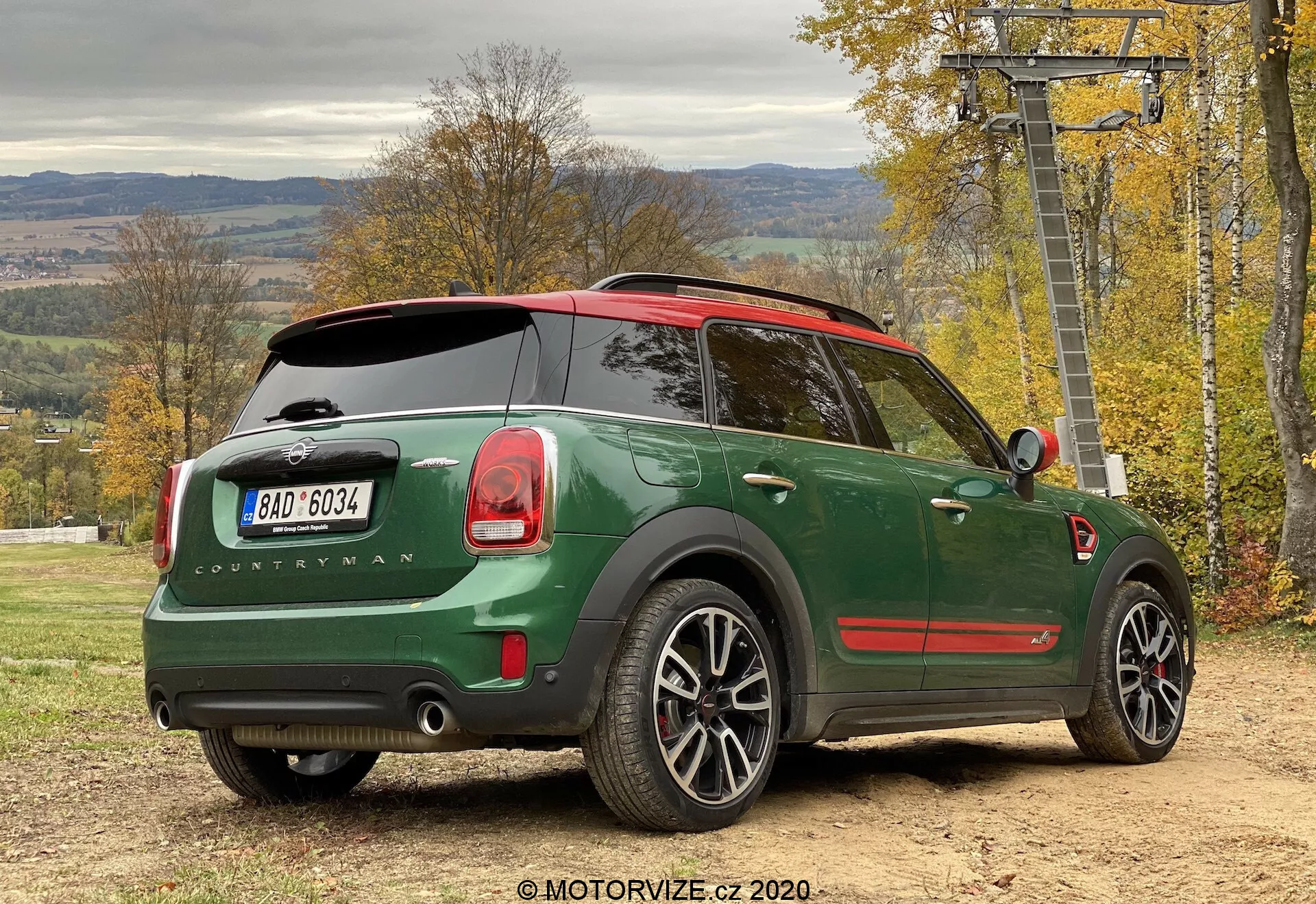 Vista posteriore tre quarti di una Mini Cooper Countryman John Cooper Works 2020 verde, parcheggiata all'aperto, che mette in risalto il design della vettura dalla prospettiva laterale posteriore. I dettagli degni di nota includono una carrozzeria verde scuro con tetto rosso, badge 'Countryman', logo Mini sul portellone posteriore, luci posteriori distintive, sistema di scarico doppio, cerchi in lega, tetto panoramico, tergicristallo posteriore, finestrini posteriori oscurati, sensori di parcheggio e specchietto laterale con indicatori di direzione integrati. Il veicolo è su una superficie non asfaltata con fogliame autunnale e una struttura di seggiovia sullo sfondo, suggerendo uno stile di vita attivo e all'aperto.