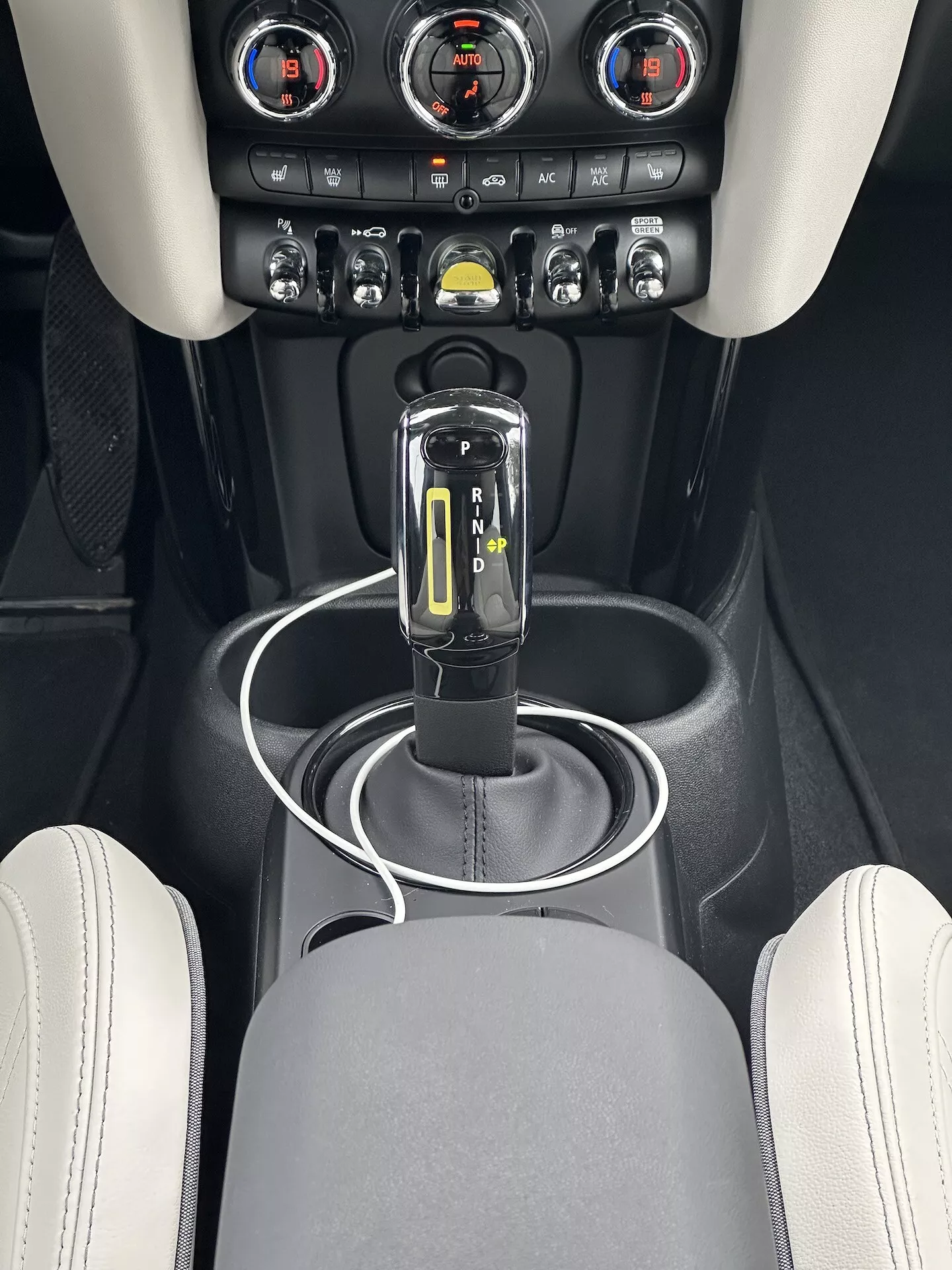Vista ravvicinata della console centrale del Mini Electric 2021, mettendo in evidenza il selettore del cambio in posizione 'P' con finitura lucida nera e cromata ed indicatori LED, circondato da una superficie nera opaca con un anello argentato, porte per la connettività USB, e area di controllo clima con manopole metalliche e display digitale della temperatura, impostato su un design interno premium con colori della tappezzeria a contrasto.