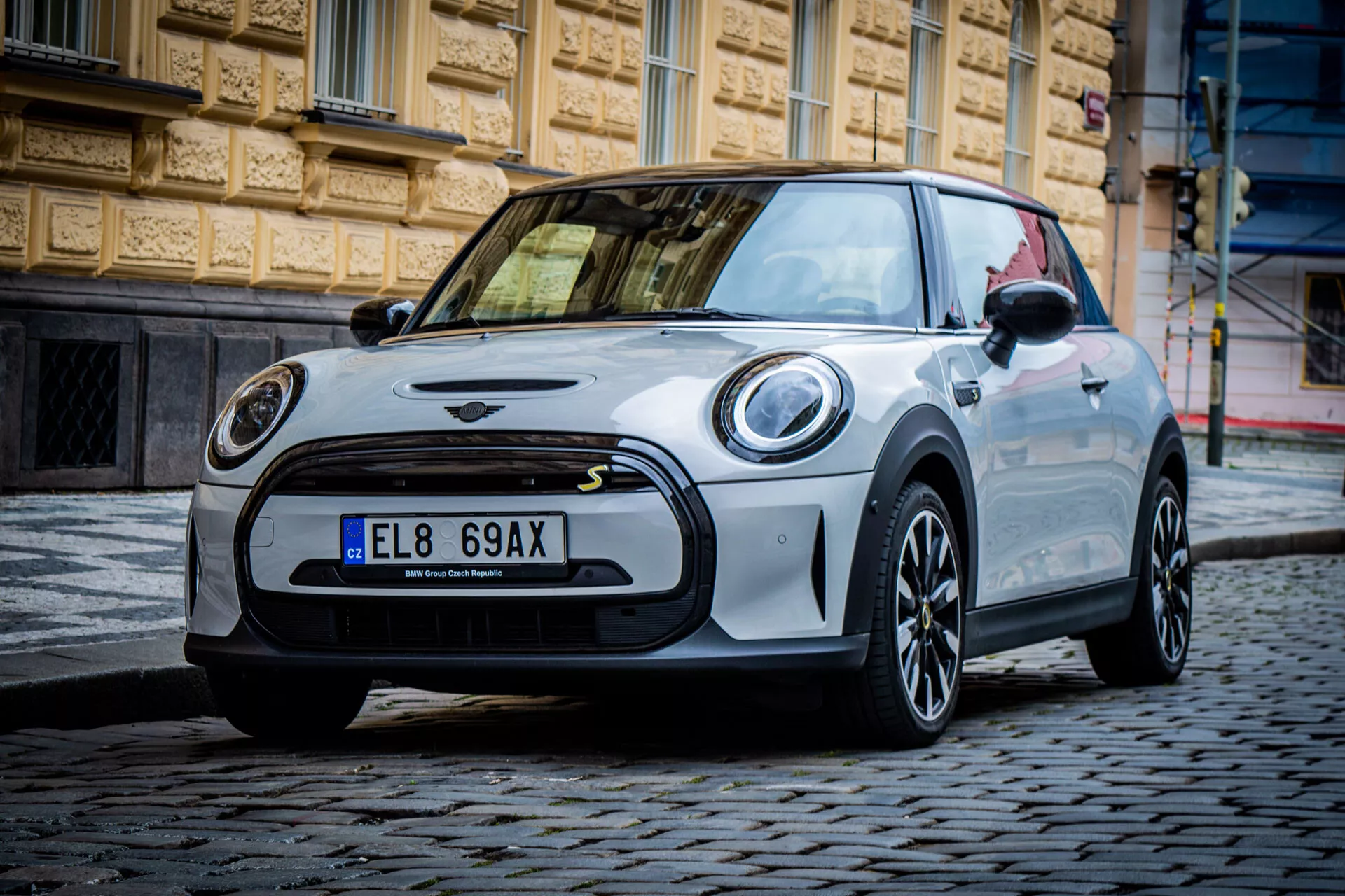 Mini Elettrica 3 porte 2021 in un contesto urbano che mostra vista frontale lato sinistro, fari LED circolari, griglia anteriore minimalista con emblema, ruote in lega scura con design a più razze, tetto e specchietti di contrasto, ed estetica sportiva su strada acciottolata