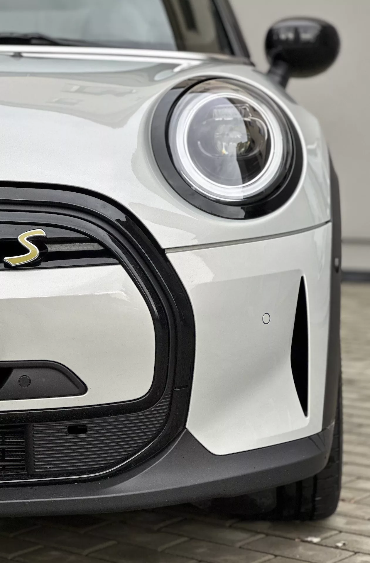 Vista ravvicinata del Mini Electric 3 porte del 2021 che mostra il lato anteriore lato guidatore, dotato di un faro circolare LED o Xenon accanto a una griglia elegante a nido d'ape con una vista parziale del distintivo sportivo, su una carrozzeria bianca solida