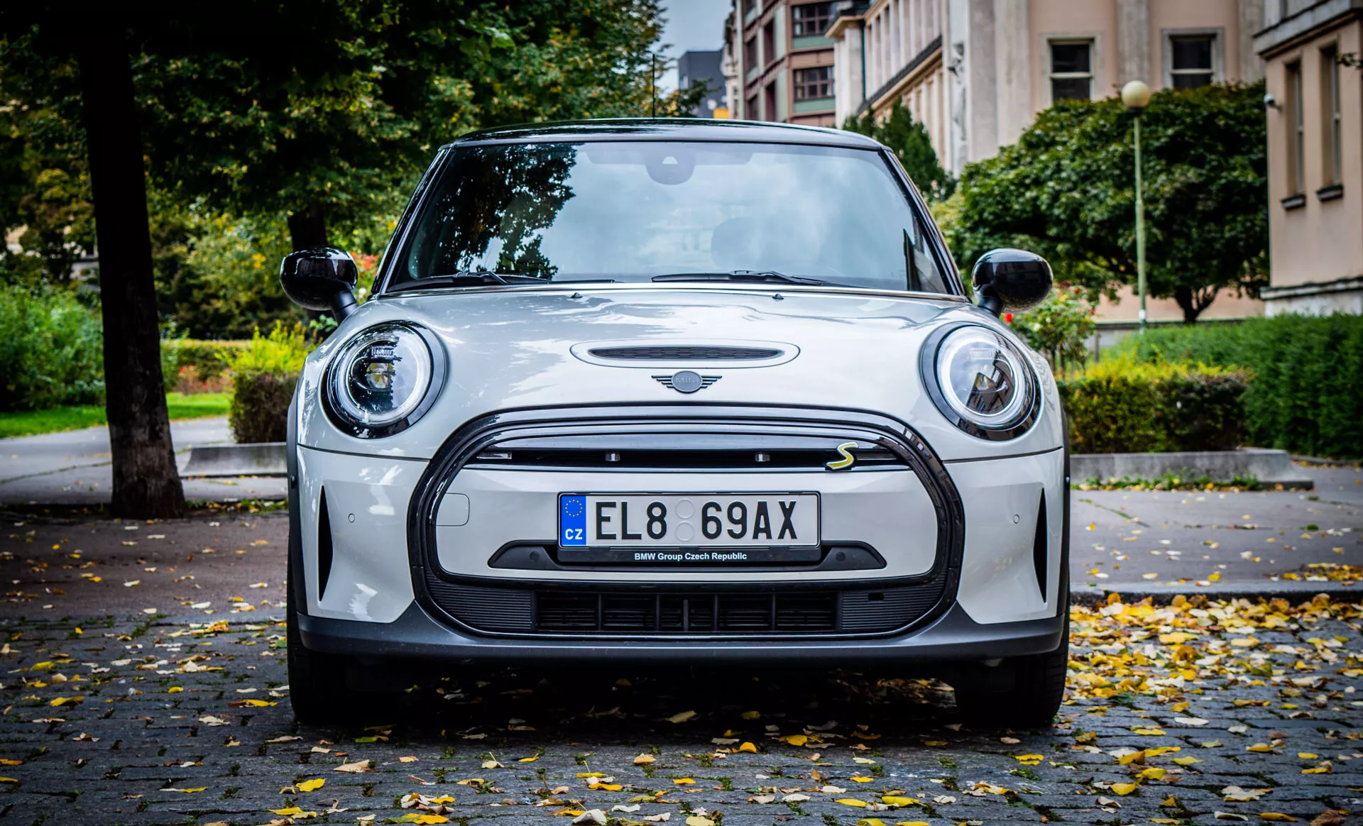 Vista frontale di una Mini Cooper 2021 Electric 3 porte su strada lastricata con foglie autunnali, che mostra tecnologia dei fari LED, sensori di assistenza al parcheggio, paraurti in stile sportivo e dettaglio dell'emblema.