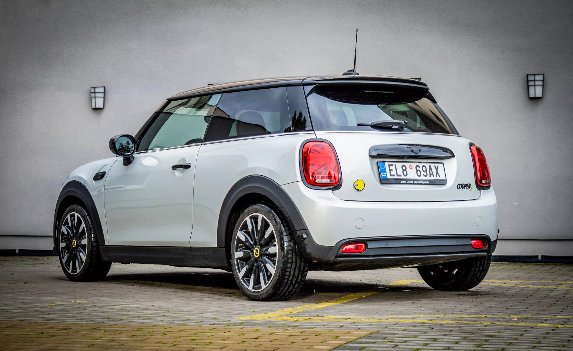 Hatchback bianca Mini Elettrica 3 Porte 2021 vista da tre quarti posteriore evidenziando i fanali posteriori circolari, l'emblema del modello, e paraurti posteriore con inserti neri, dotata di tergicristallo posteriore e spoiler sul tetto, profilo laterale mostra linee aerodinamiche, ruote nere a più razze, maniglie delle porte cromate e specchietti con indicatori di direzione integrati su sfondo neutro