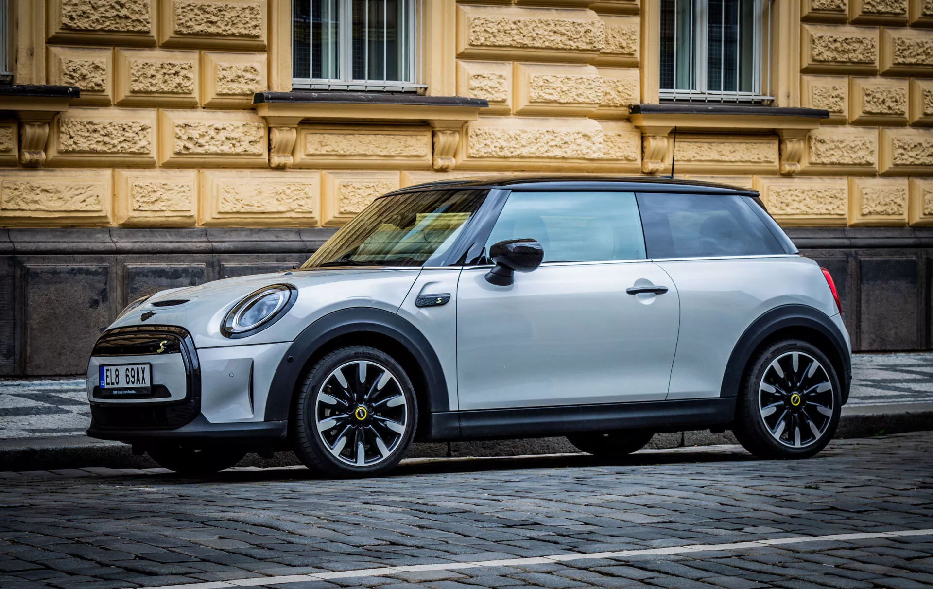 Mini Cooper elettrica a 3 porte del 2021 parcheggiata su una strada lastricata con un edificio storico ornato sullo sfondo, che mostra il suo design moderno e le caratteristiche ecocompatibili