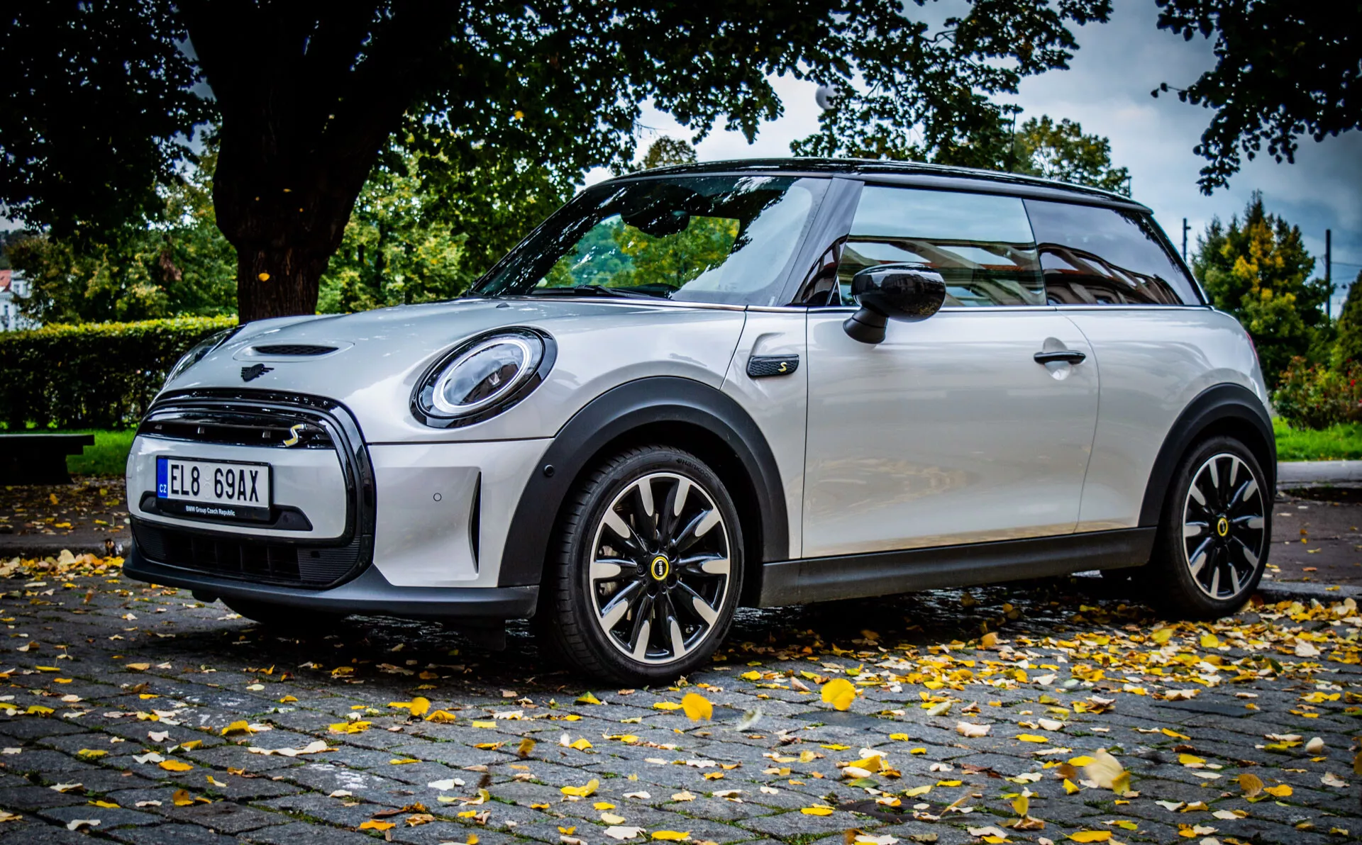 Mini Elettrica 3 porte del 2021 in schema di colori bicolore argento e nero, ripresa da un angolo basso dinamico che evidenzia il suo design sportivo, fari anteriori circolari LED e cerchi in lega neri con dettagli a contrasto, parcheggiata su una strada urbana.