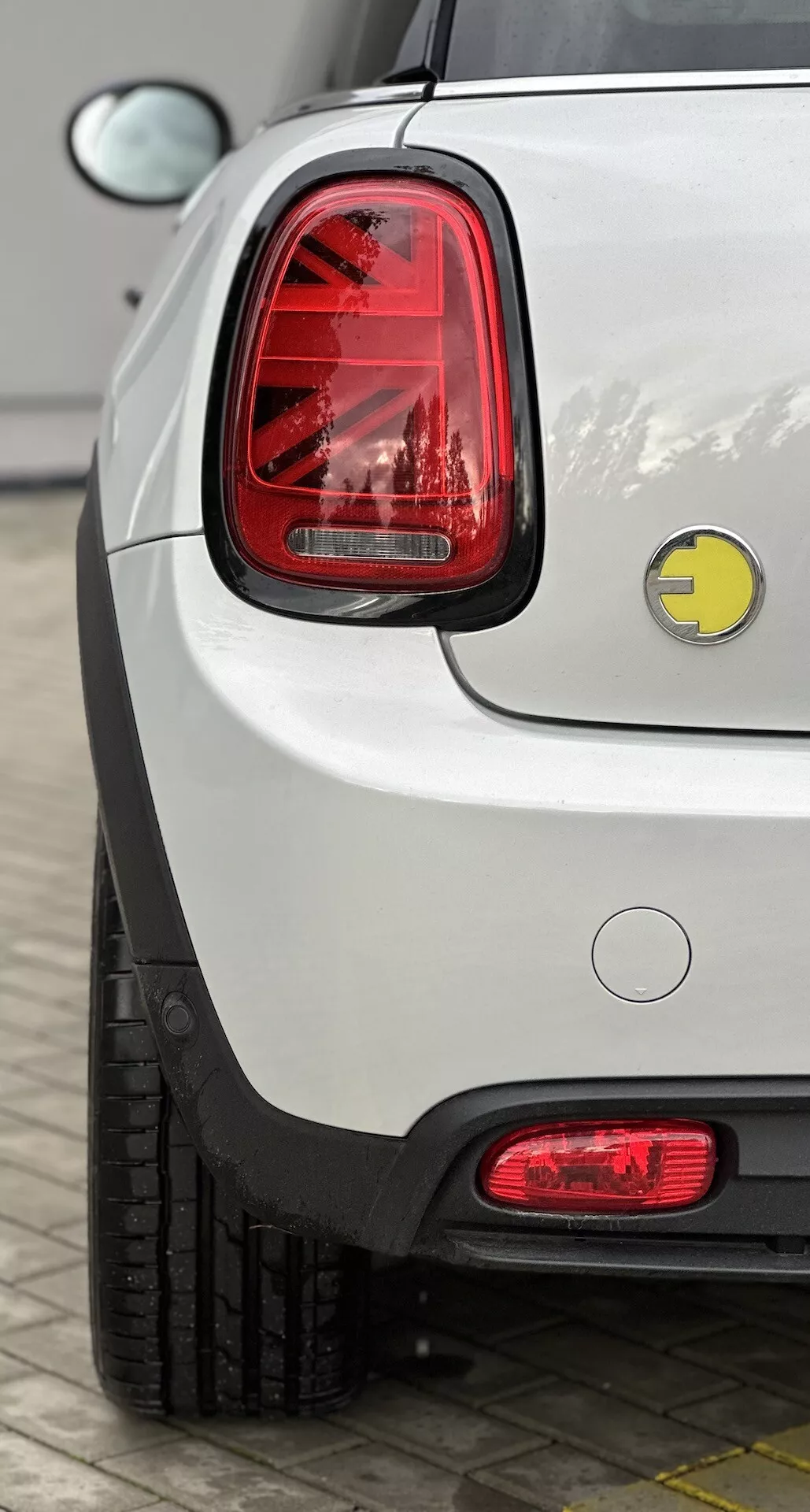 Vista ravvicinata dell'angolo posteriore destro di una Mini Electric 2021 bianca, che evidenzia la sua finitura lucida, le luci posteriori LED verticali, il badge del marchio, il tappo del carburante convenzionale, il paraurti posteriore con riflettore, i sensori di parcheggio e la sezione del pneumatico visibile, parcheggiata all'esterno.
