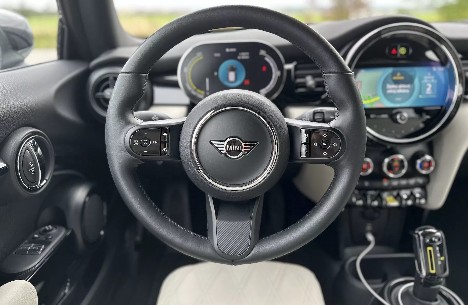 Vista interna della Mini Electric 3-Porte 2021 dal punto di vista del conducente, con volante multifunzione con emblema Mini Cooper, cruscotto digitale, sistema di infotainment con schermo touchscreen centrale, leva del cambio automatico e sedili dal design bicolore.