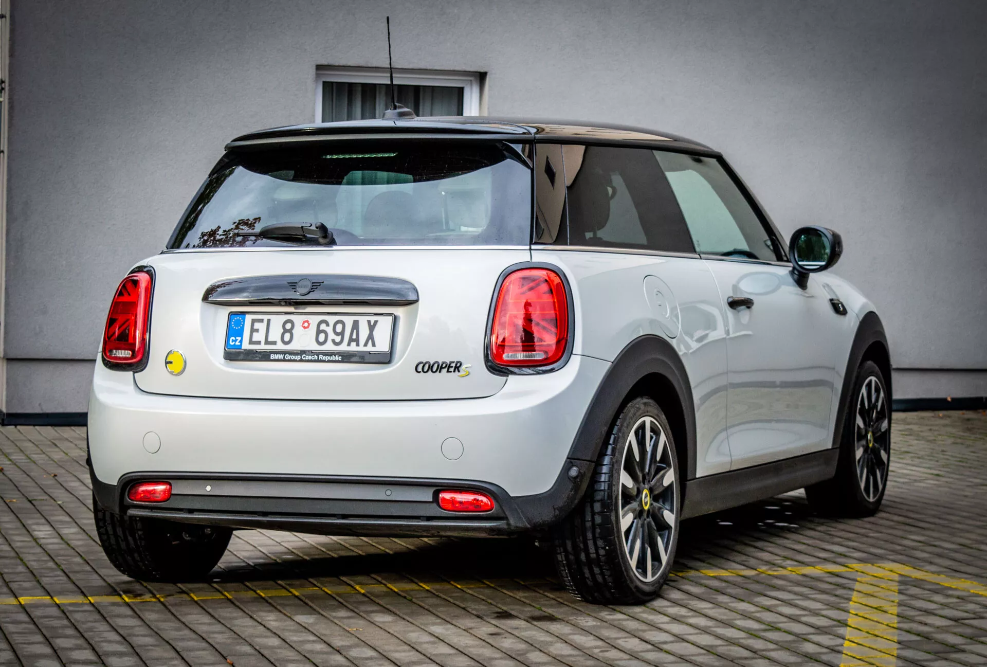 Vista posteriore laterale di una Mini Electric 3-Door Cooper bianca 2021, che mette in mostra il suo design distintivo delle ruote e le iconiche luci posteriori cerchiate in cromo, parcheggiata davanti a un edificio commerciale