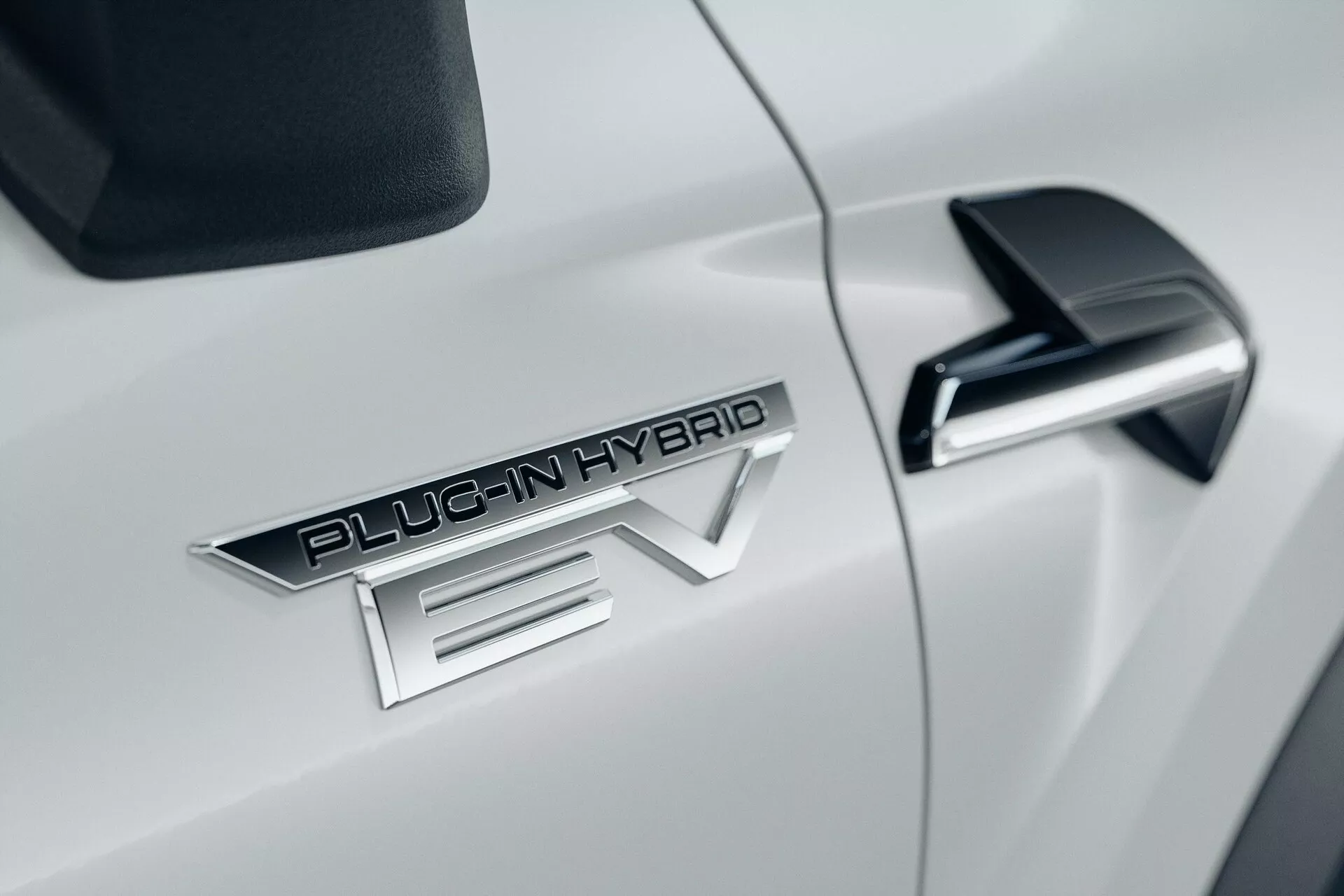 Primo piano del lato dell'auto bianca Mitsubishi ASX 2023 di seconda generazione, con il badge prominente 'PLUG-IN HYBRID EV', elegante specchietto laterale nero con freccia integrata e carrozzeria aerodinamica bianca lucida.
